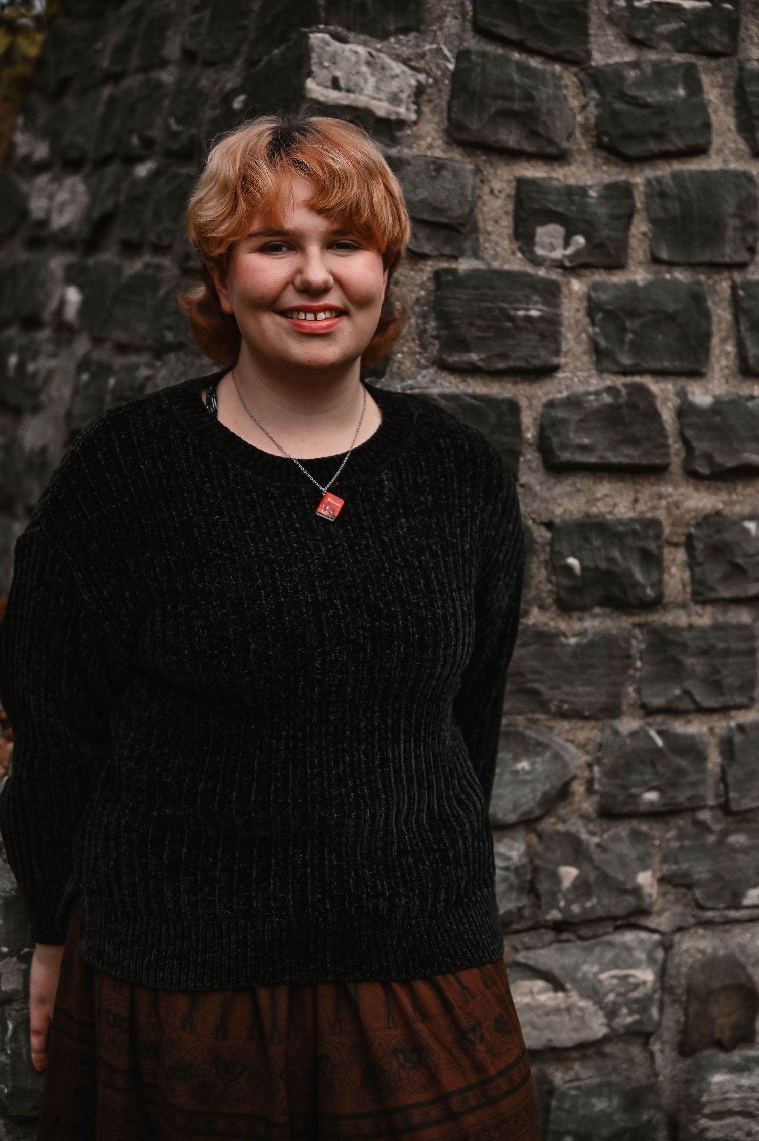 Portrait series against rustic stone wall wearing black sweater and brown skirt.