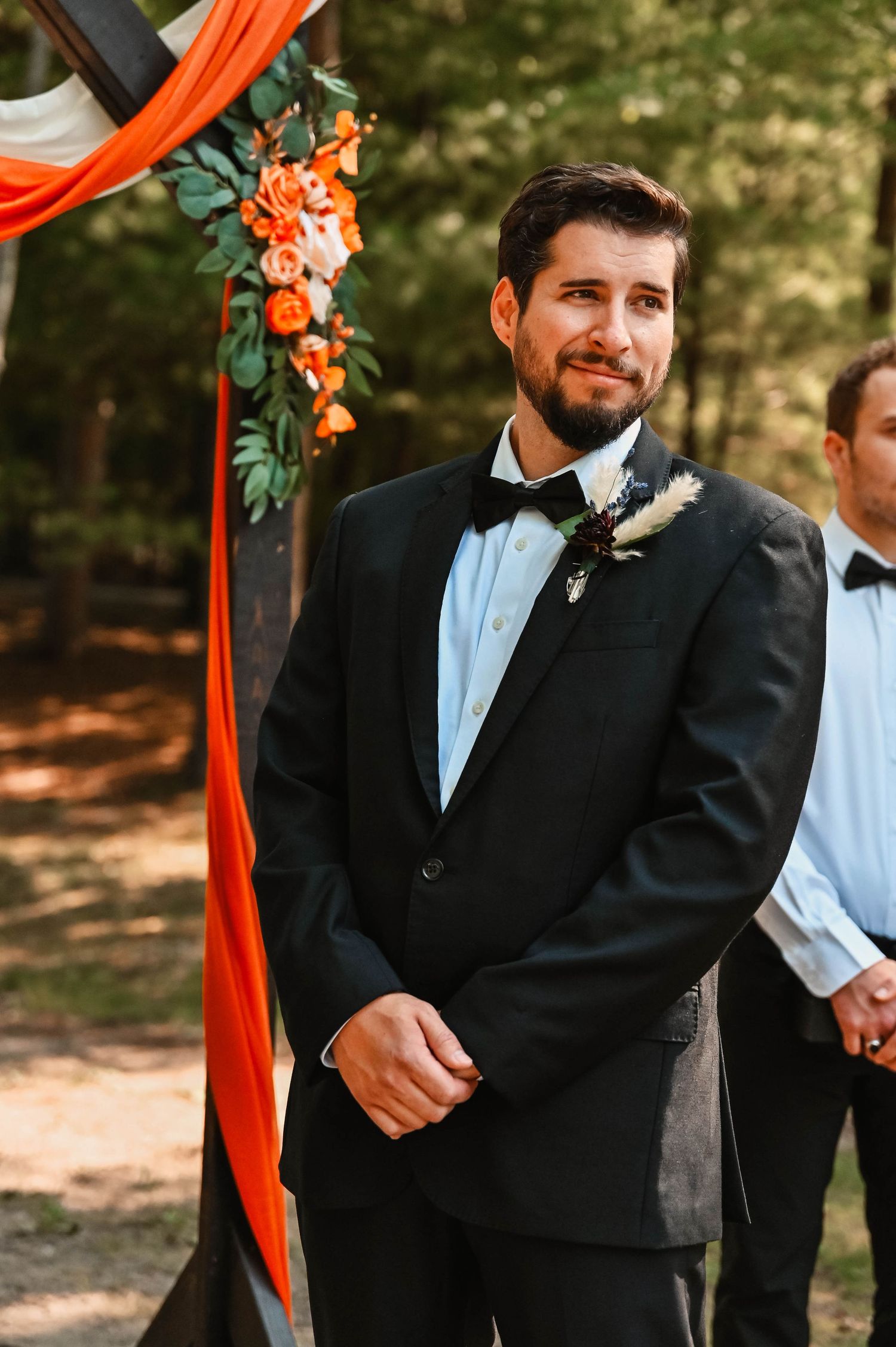 Groom stands under orange floral arch during outdoor forest wedding ceremony.
