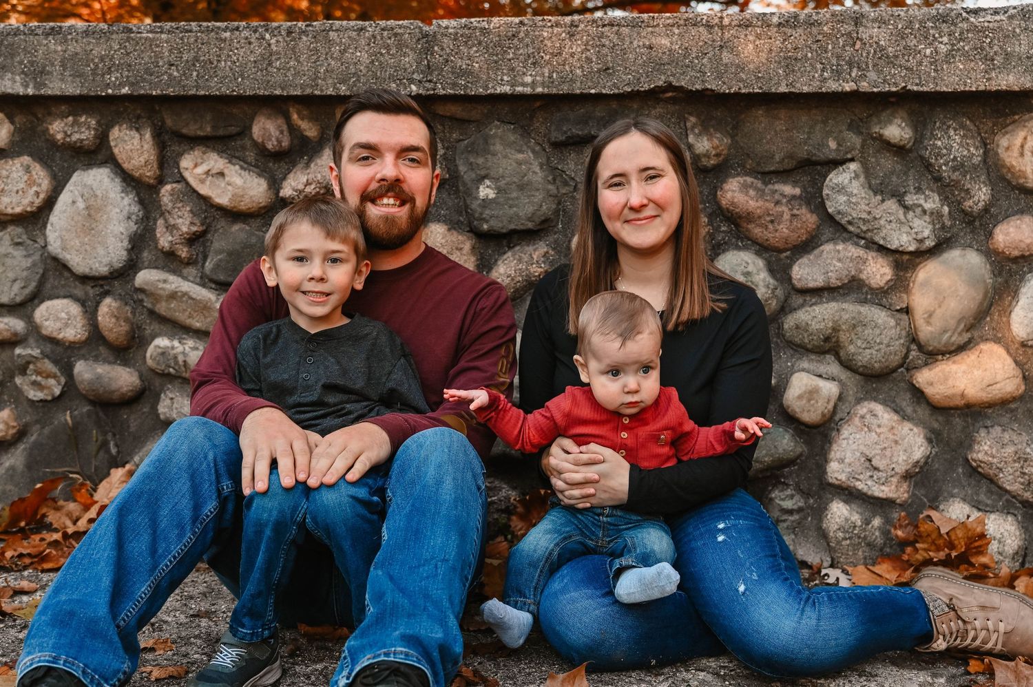 Family of four sits casually on stone wall during autumn photo session.