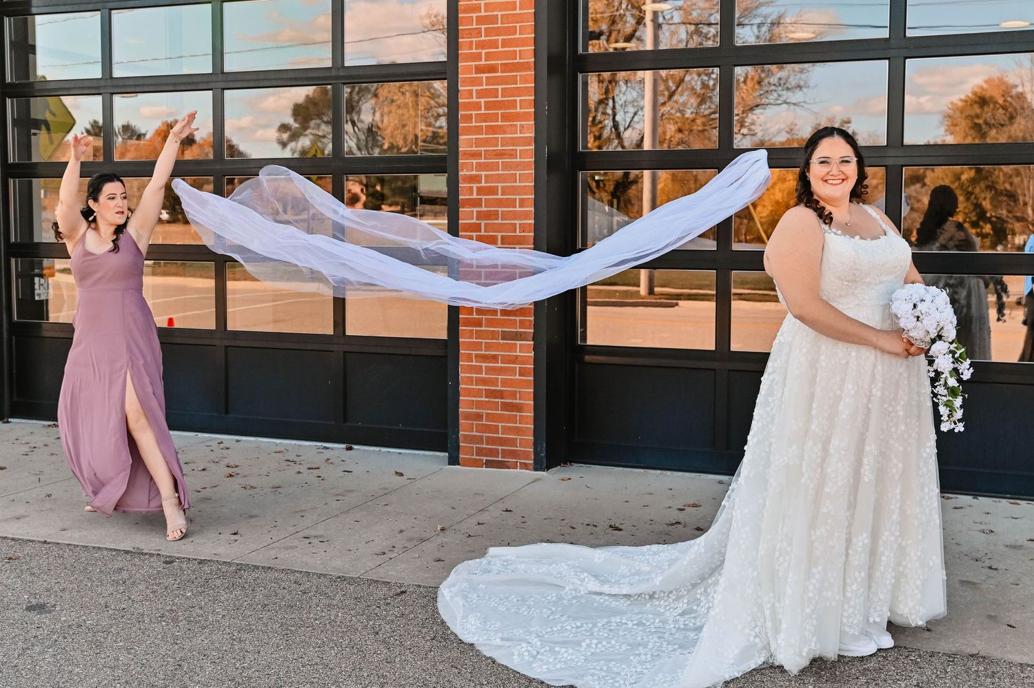 A wedding photo showing a flowing veil being tossed in the wind against a brick building with large windows.