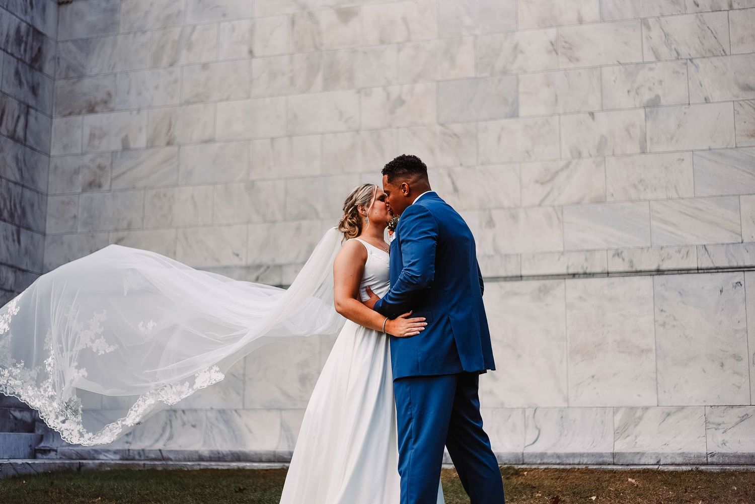 Newlywed couple embracing with flowing veil against marble wall backdrop.