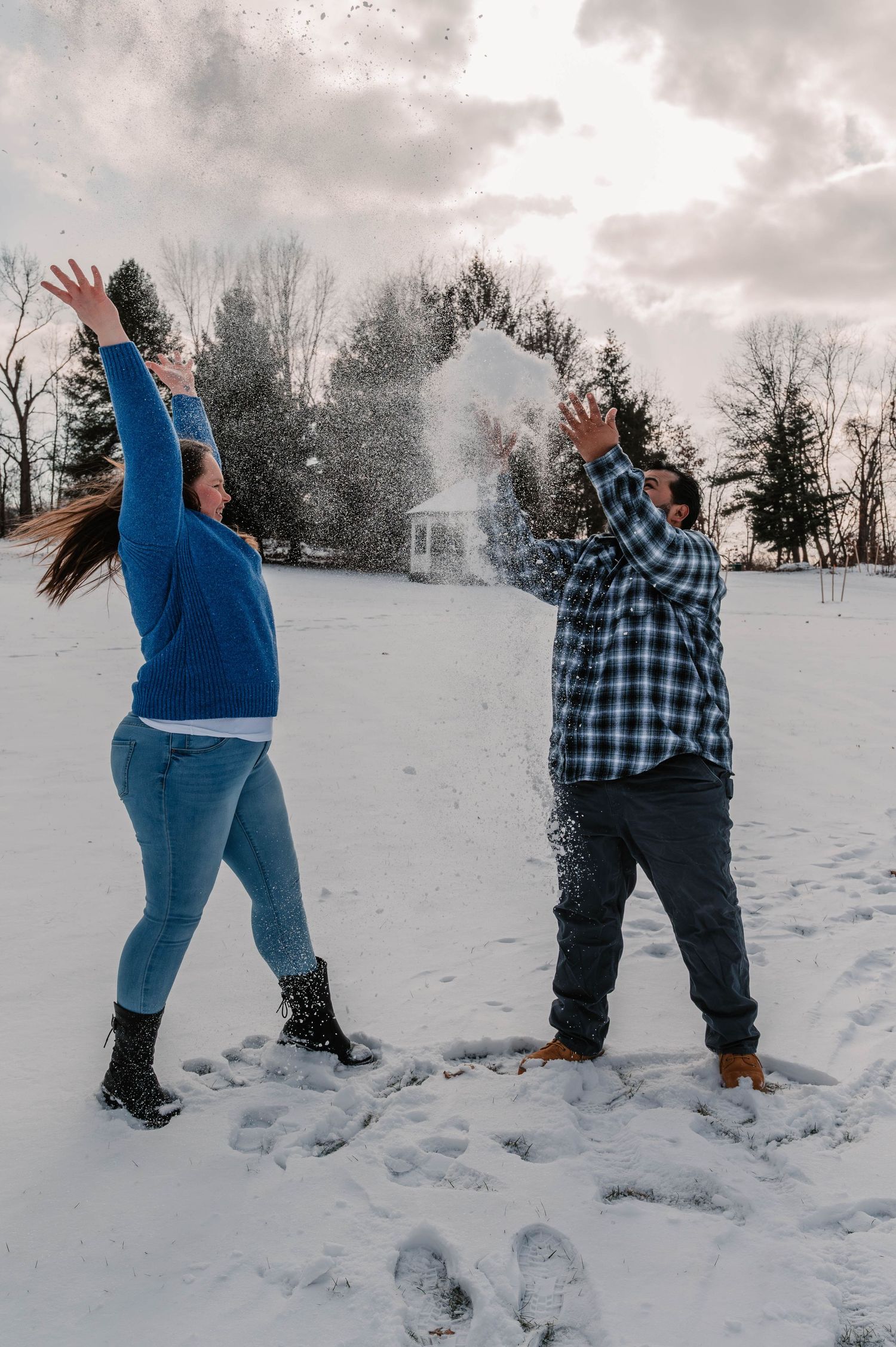 Two people throwing snow at each other in a wintry field with evergreen trees.