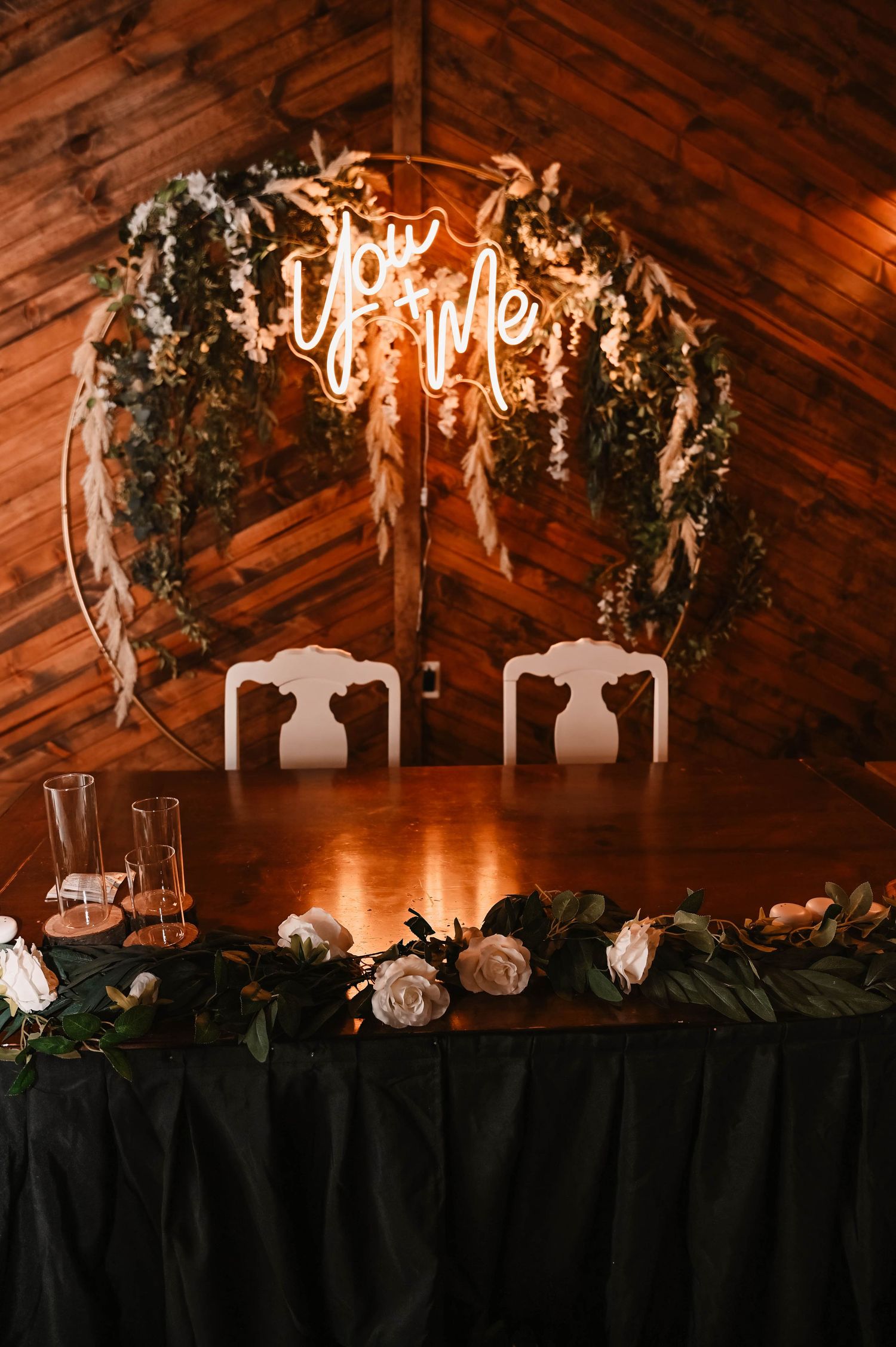 Romantic sweetheart table setup with neon sign and floral arch against wooden wall.