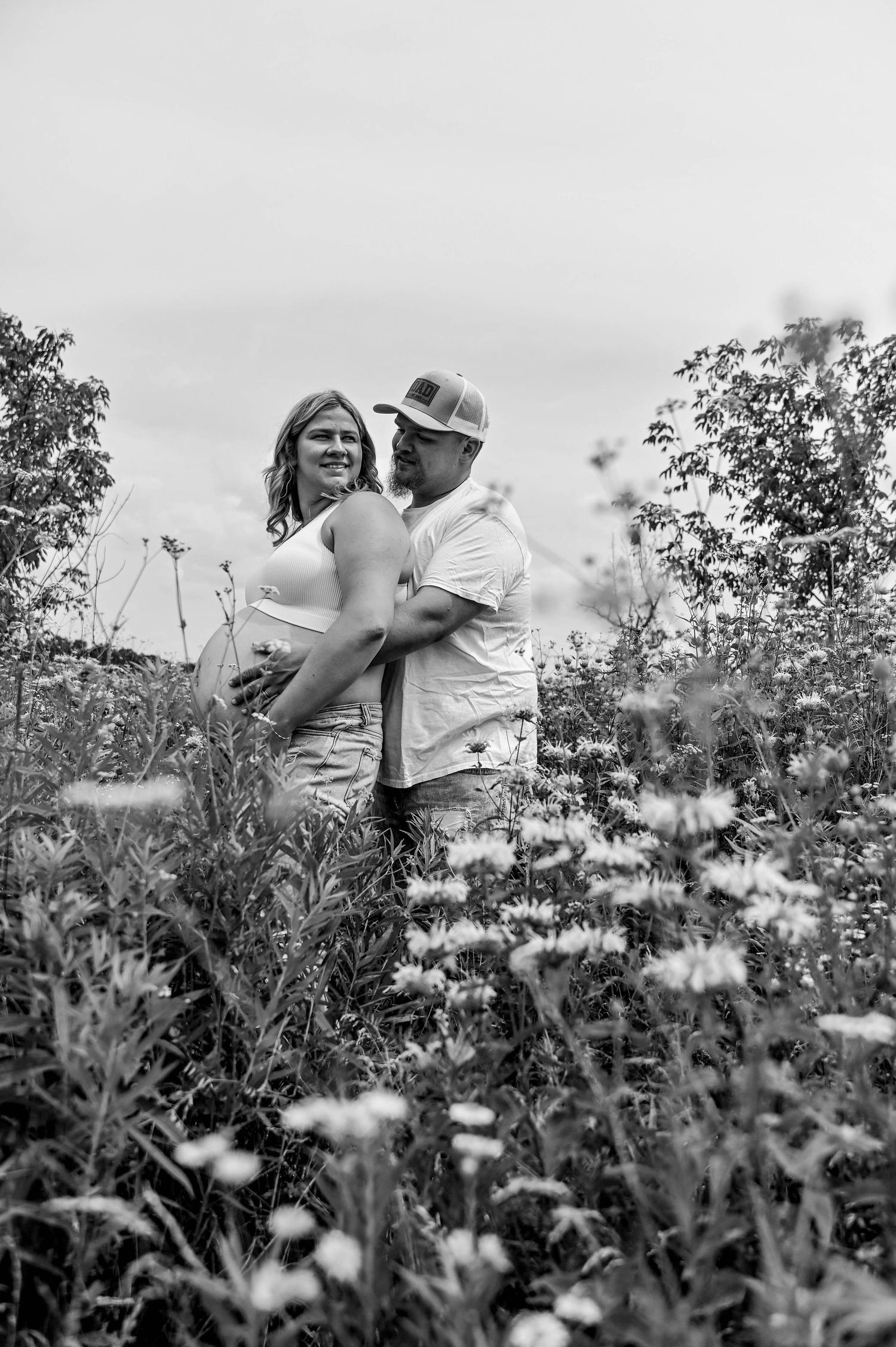 A black and white maternity photo of a couple embracing in a daisy field.