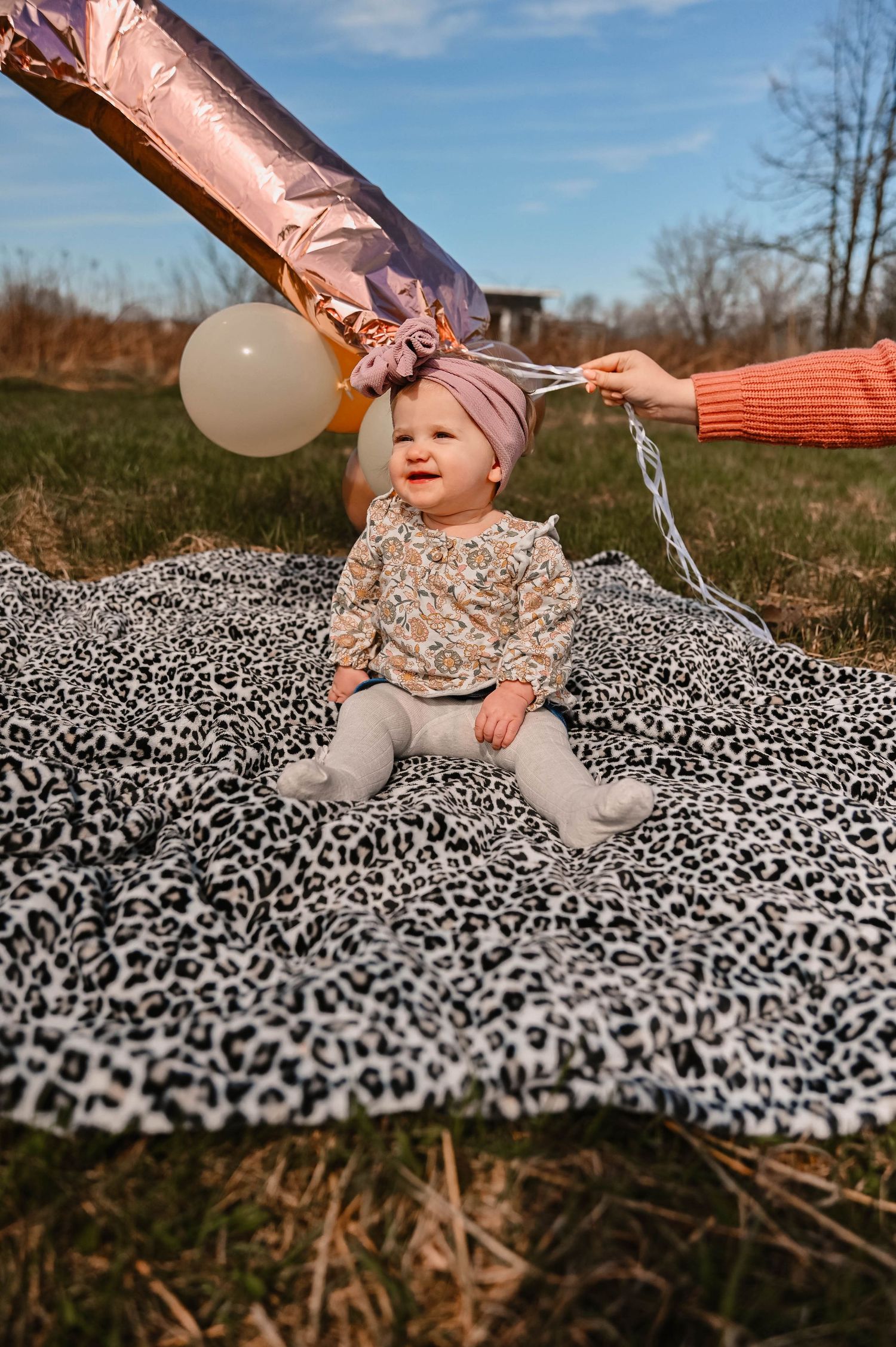 Baby in sparkly outfit sits on leopard print blanket outdoors with pink balloons.