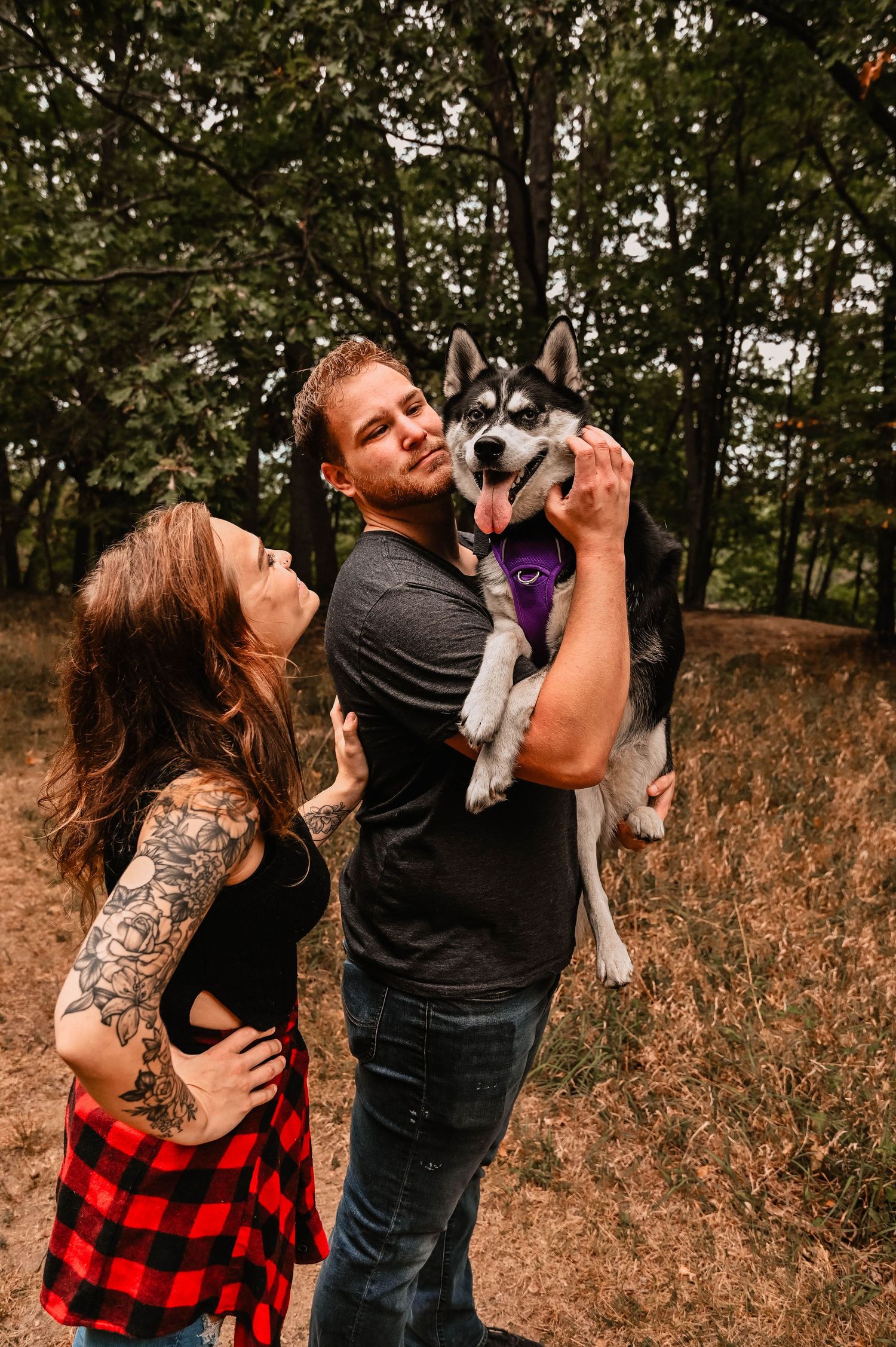 A couple poses with their husky dog in a wooded area during autumn.