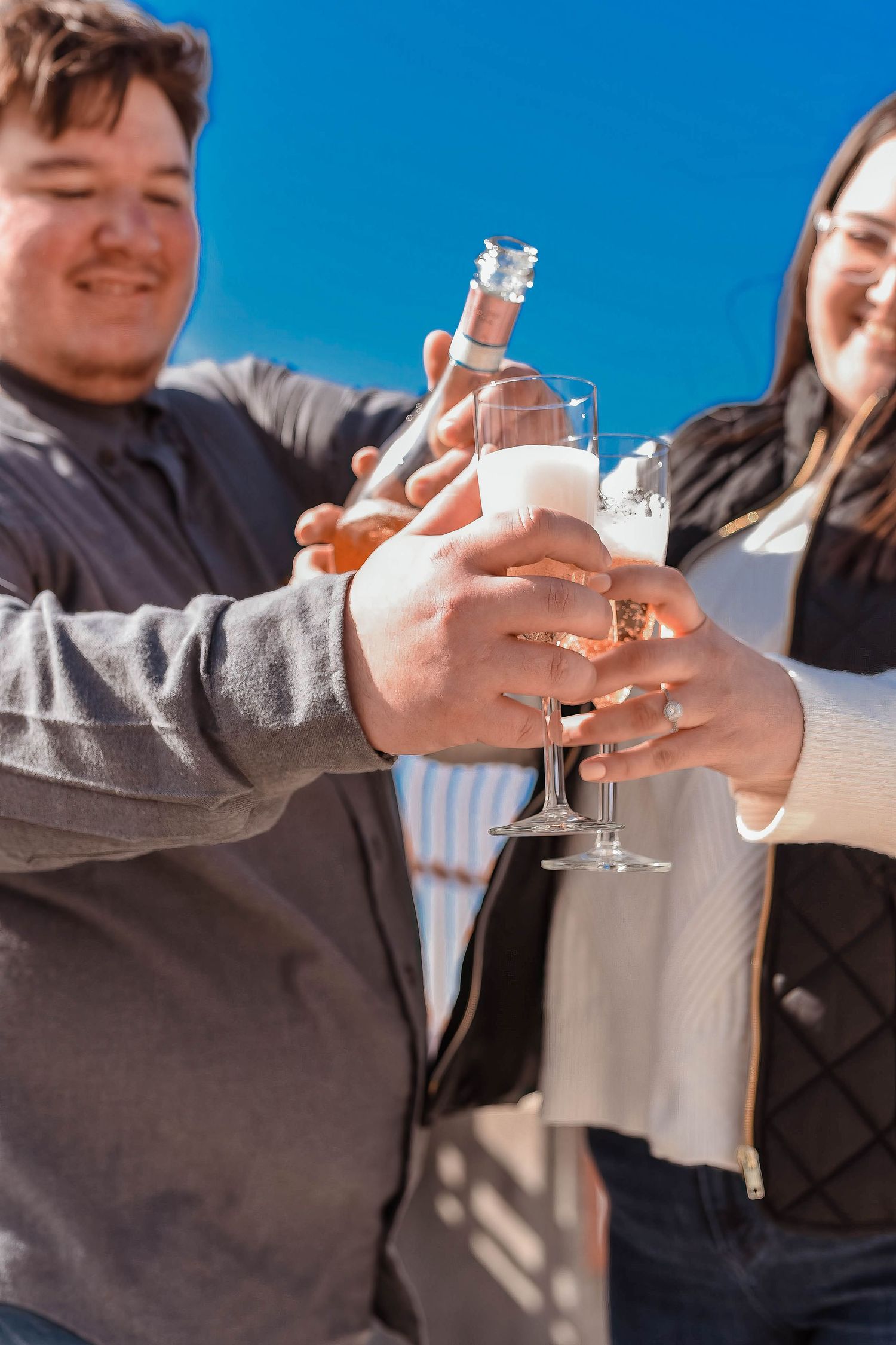 A couple toasts with champagne glasses against a bright blue sky background.