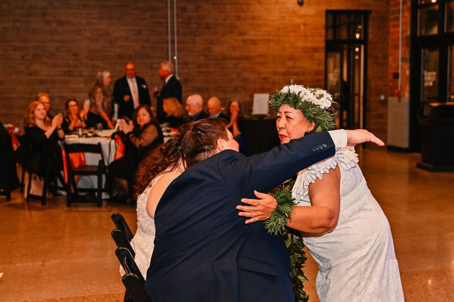 Wedding guests dance together on a wooden floor under warm lighting at an indoor reception venue.