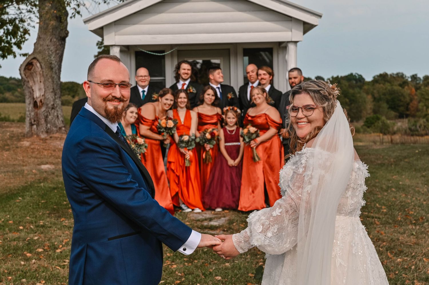 Wedding party in coral dresses and suits posing outdoors near a white building.