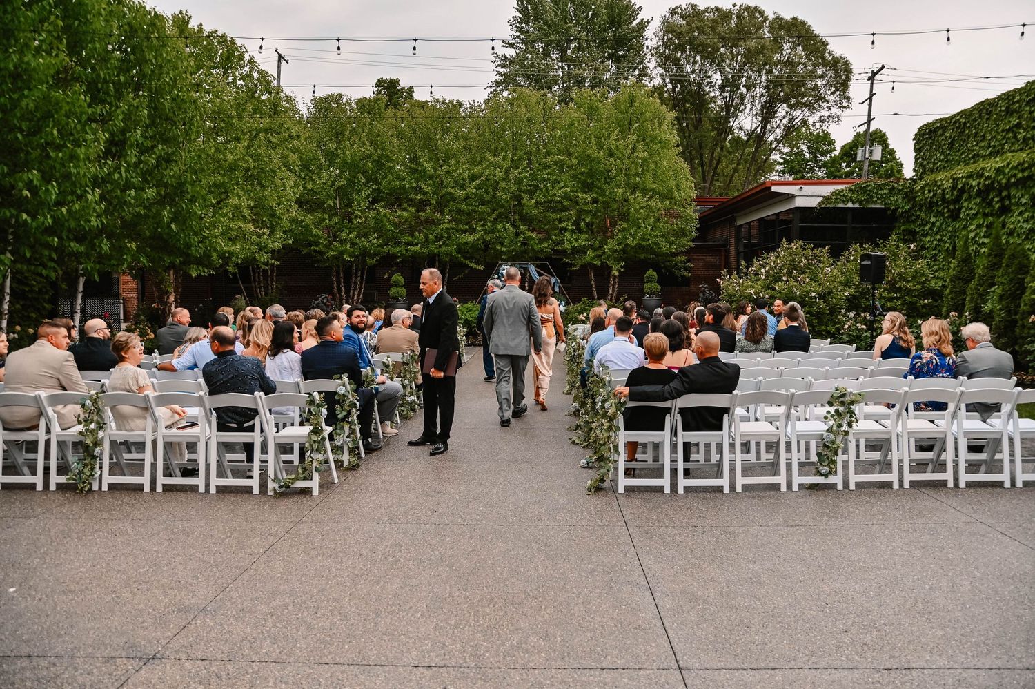 Outdoor wedding ceremony setup with white chairs and string lights under trees.