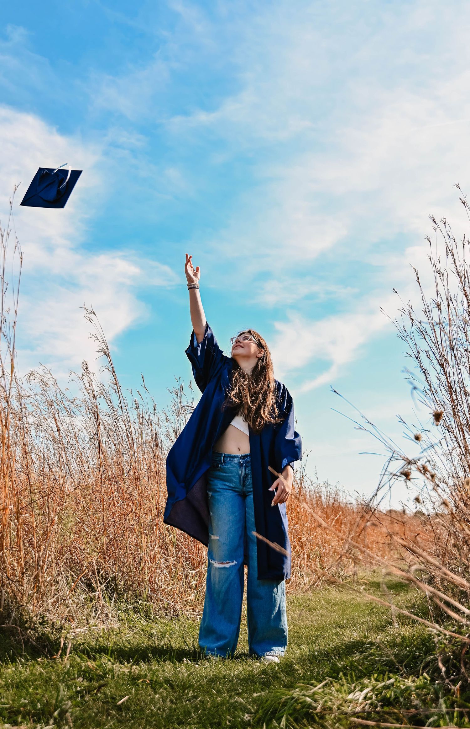 Graduate throwing cap in celebration among autumn grass under bright blue sky.