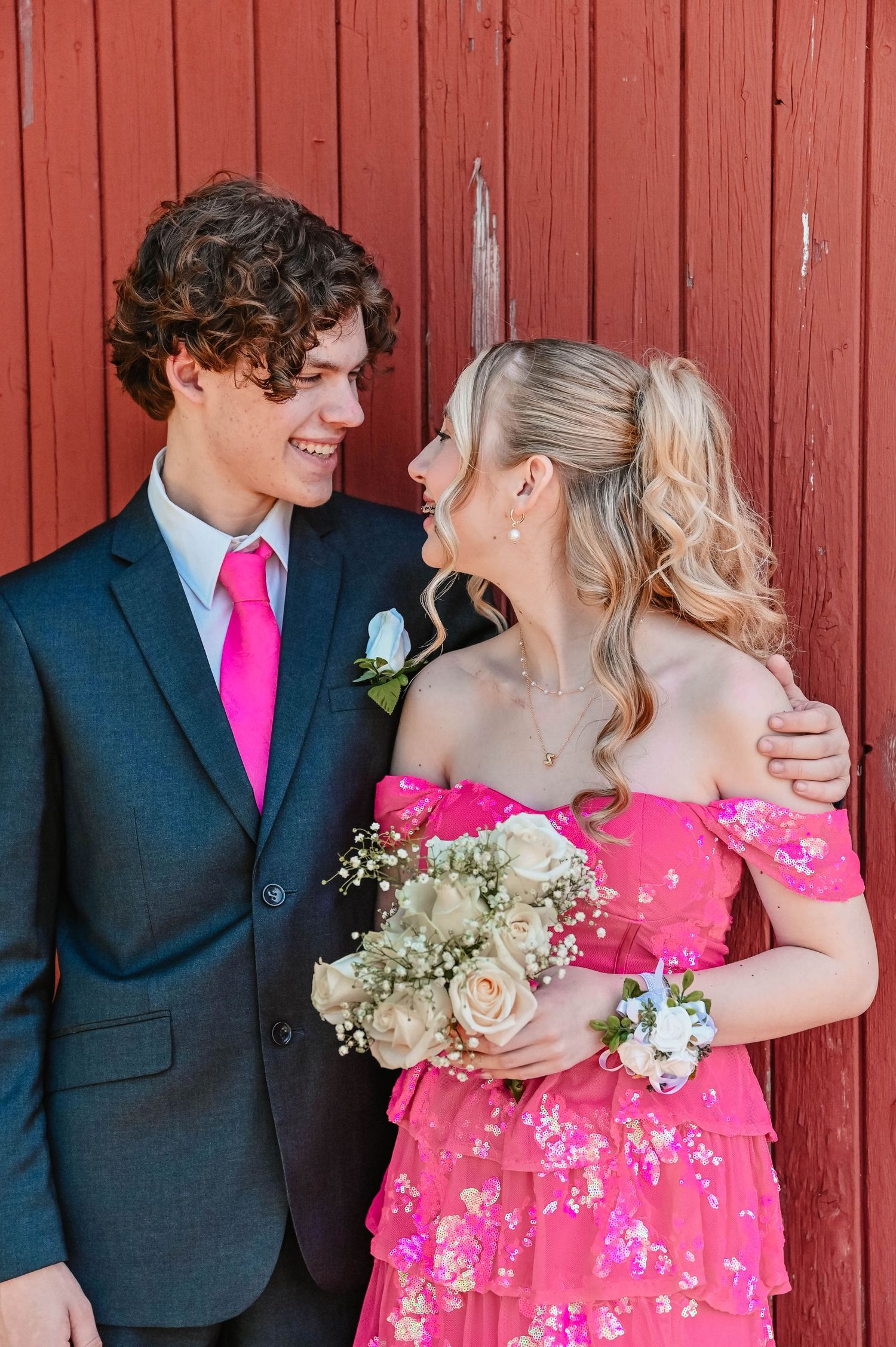 Wedding couple poses playfully against red barn wall with bride in bright pink floral dress.