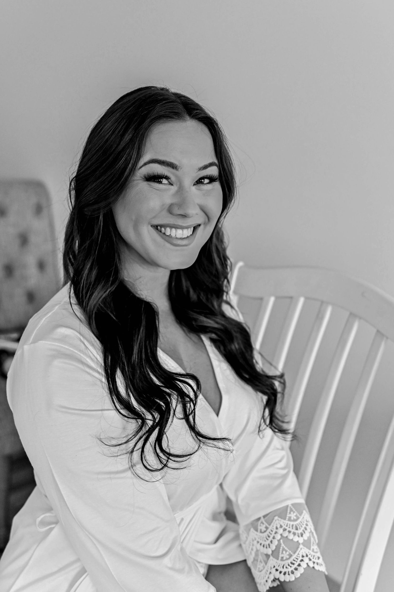 A black and white portrait of someone wearing a white lace-trimmed robe while sitting in a wooden chair.