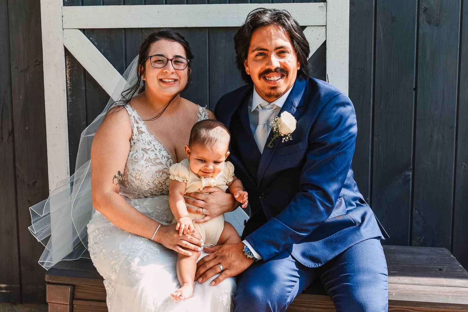 A couple in wedding attire poses with their baby in front of a rustic barn backdrop.