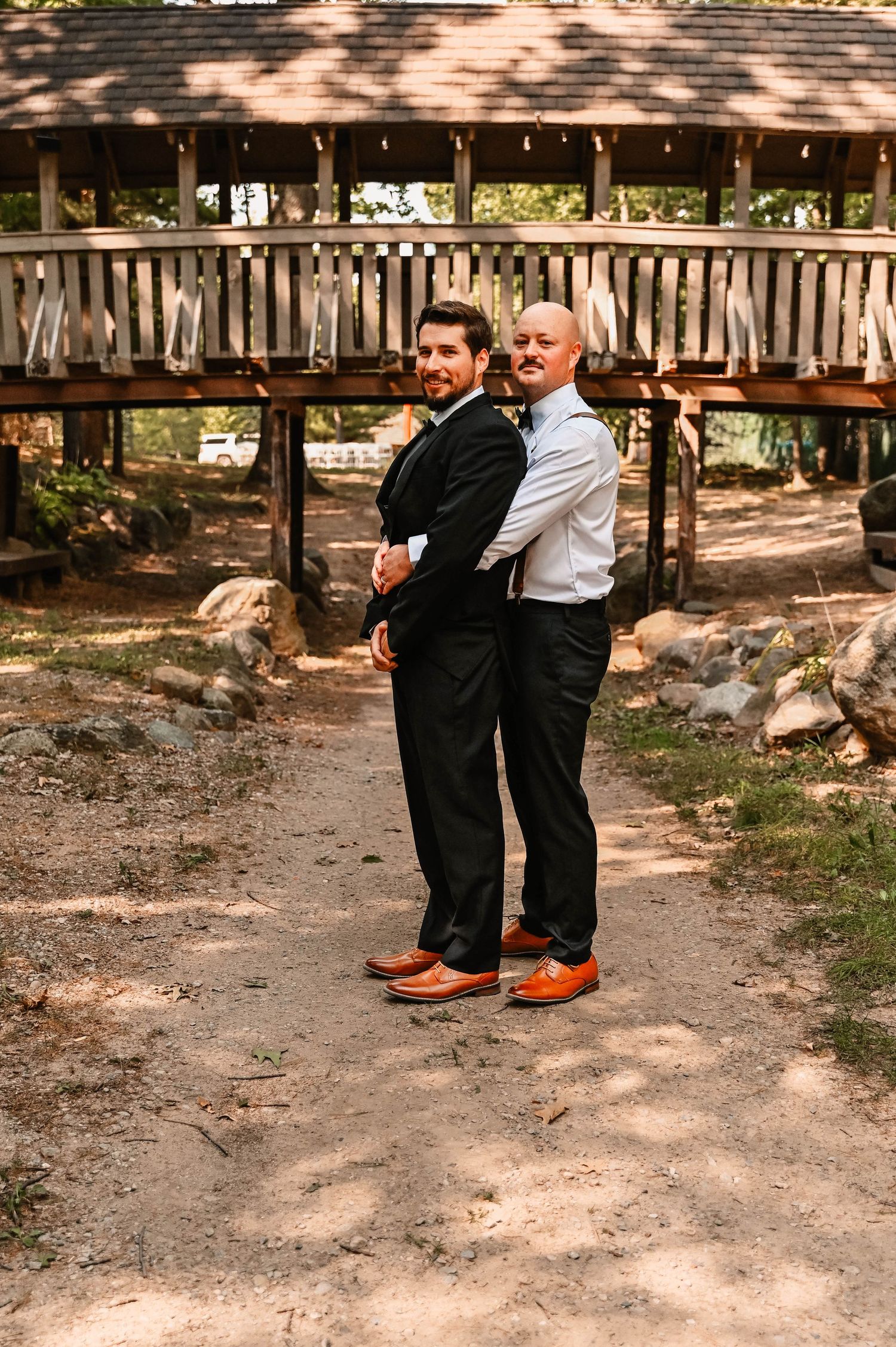 Two people pose back-to-back in formal attire near rustic wooden bridge.