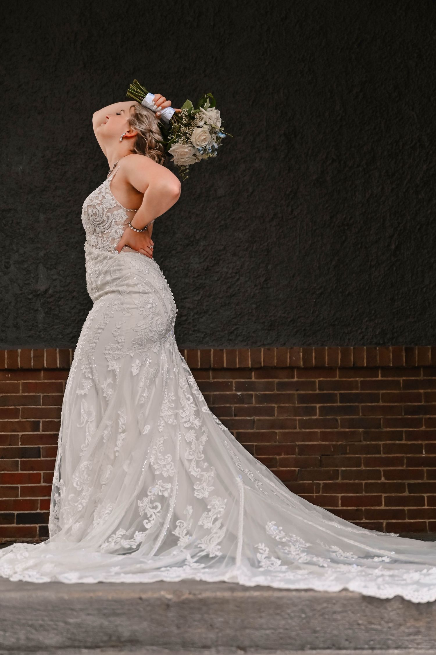 Bride in lace wedding gown with train poses against brick wall holding bouquet.