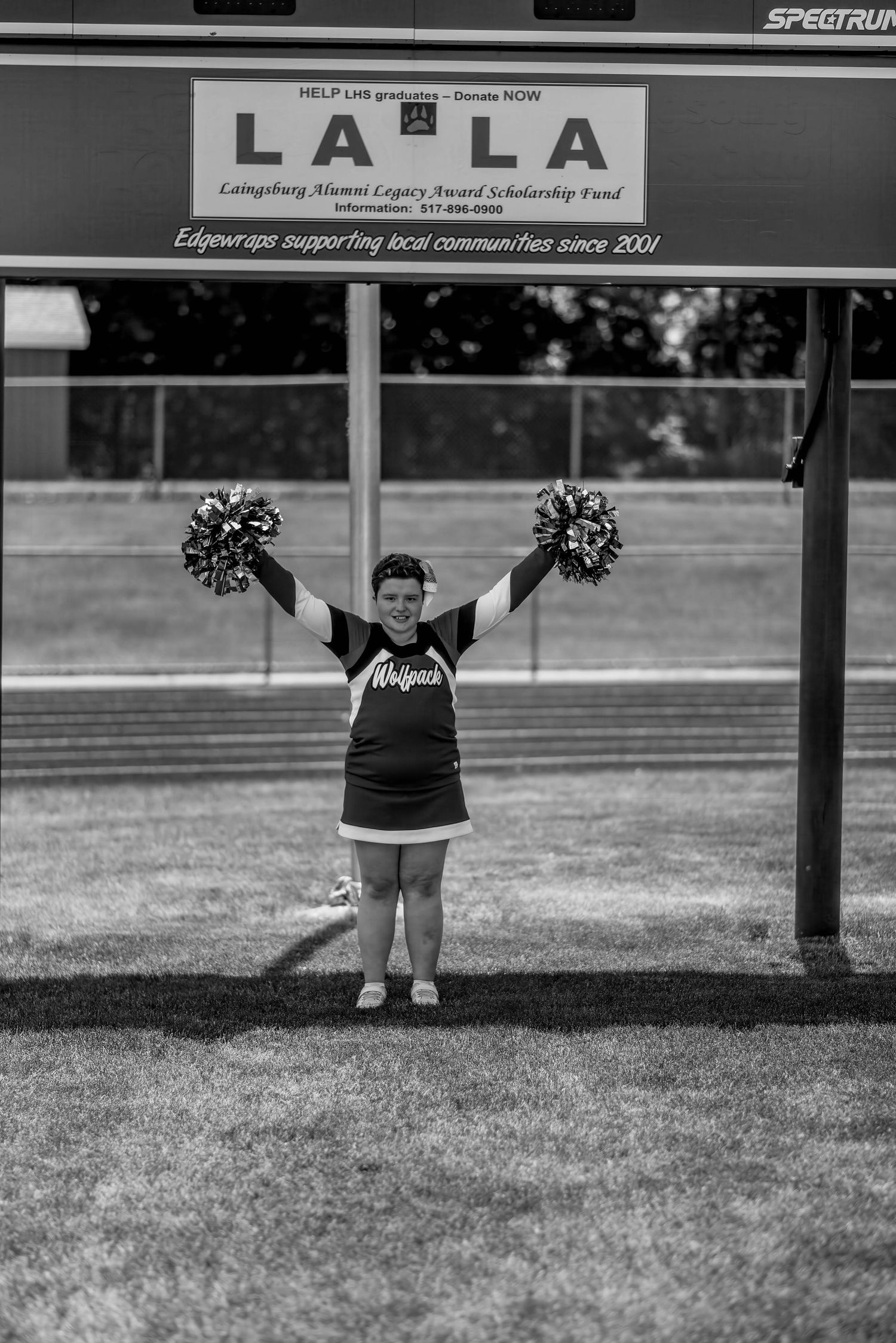 A cheerleader poses with pom poms under a LLLA sign at a sports field.