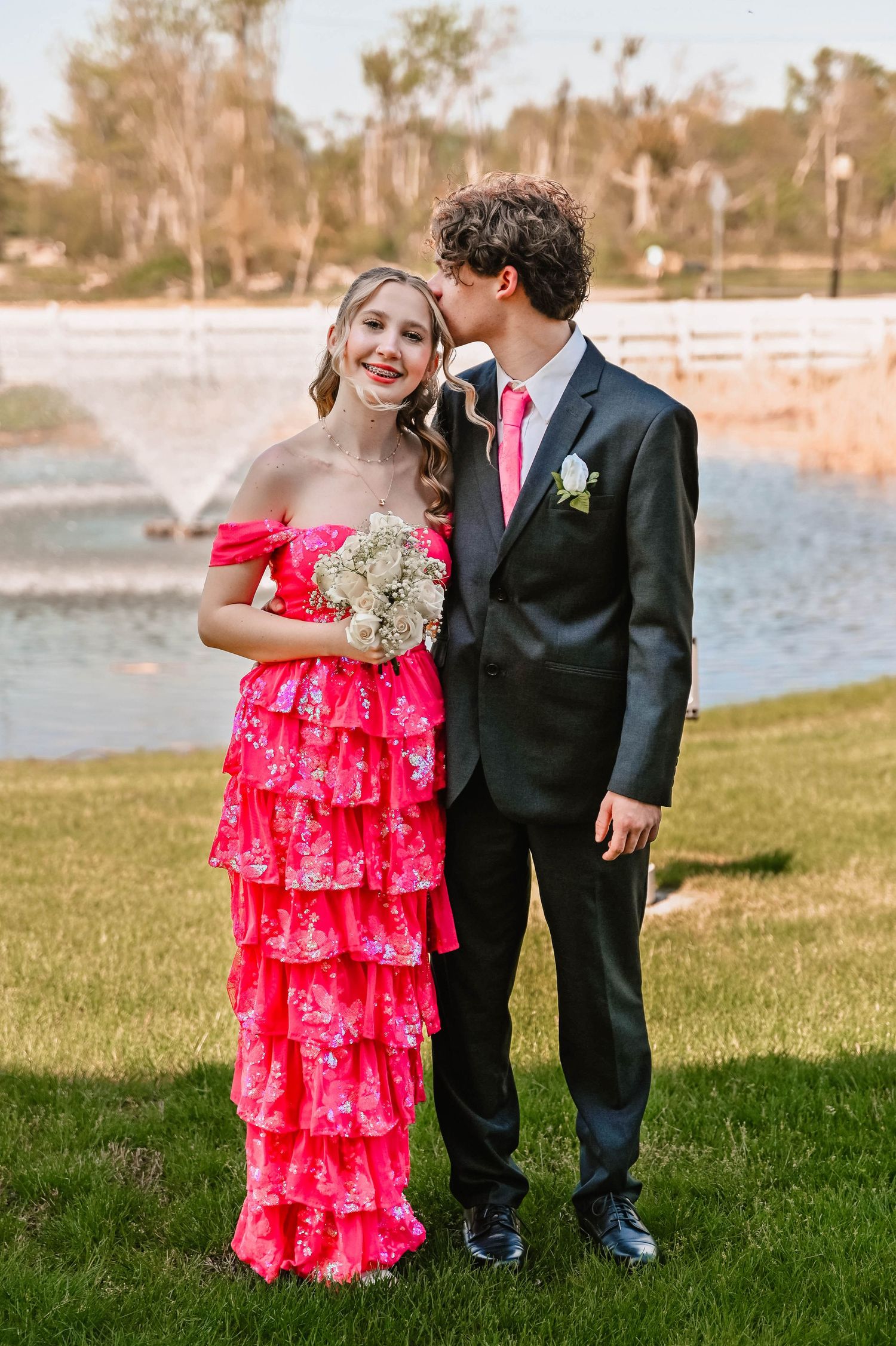 Couple in formal wear stands by a lake with vibrant pink ruffled dress and black suit.