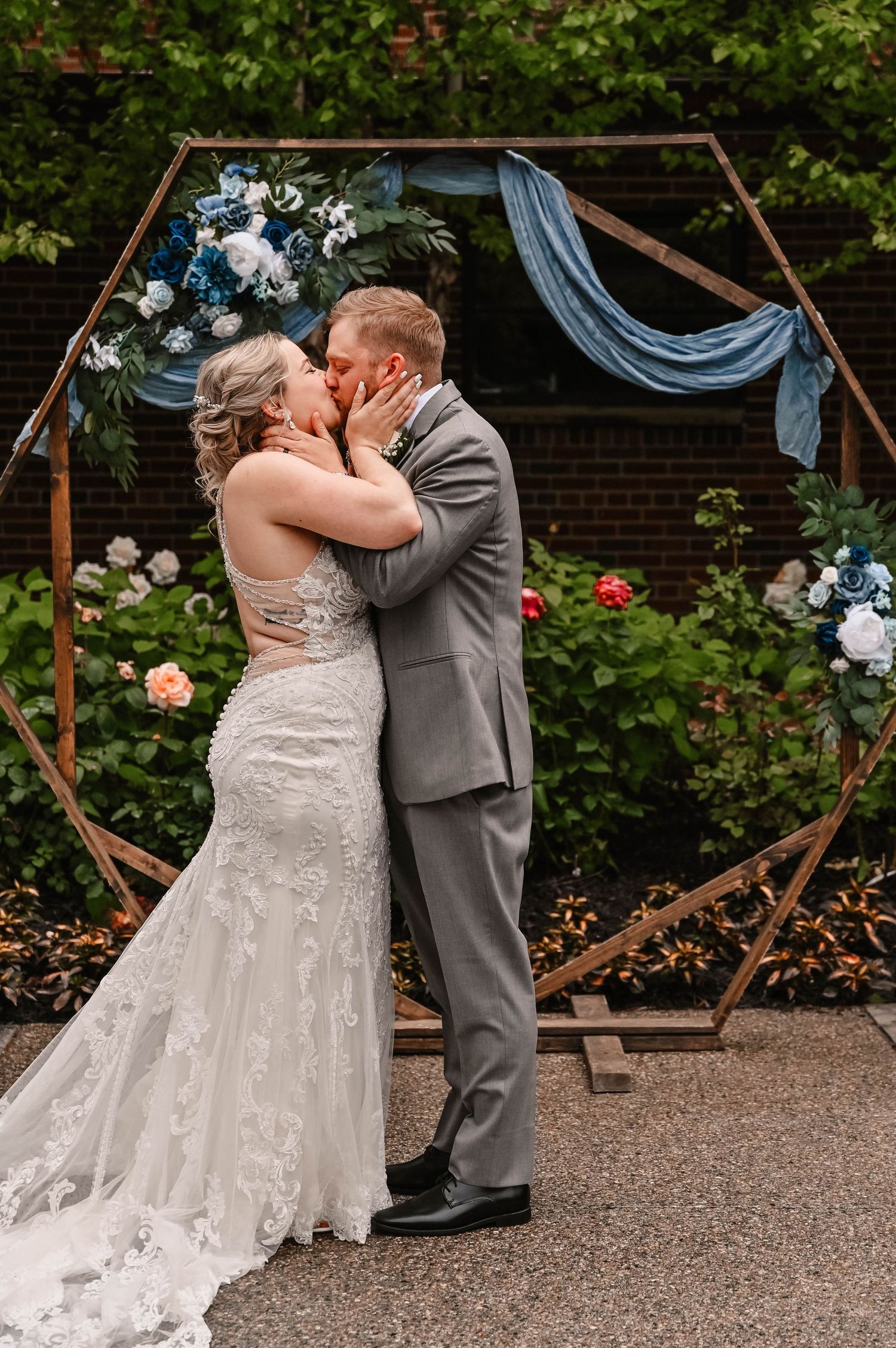 Couple shares romantic kiss under hexagonal arch decorated with blue fabric and white flowers.