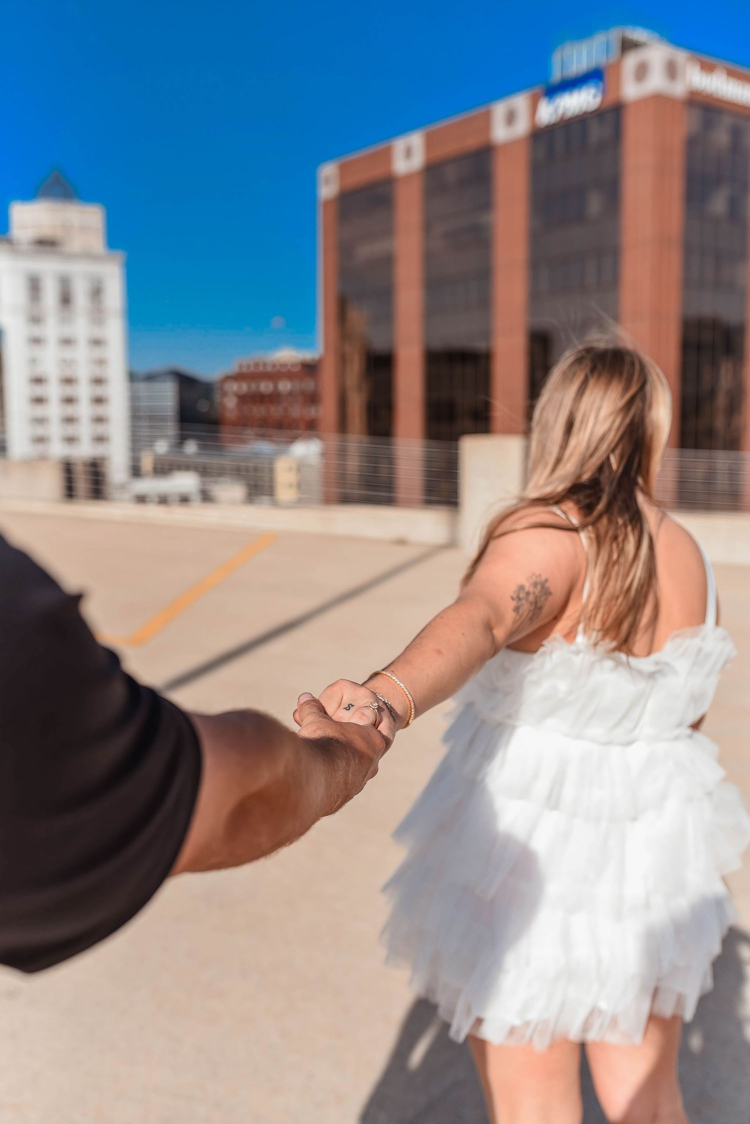 A couple poses against an urban backdrop with modern buildings and blue sky.