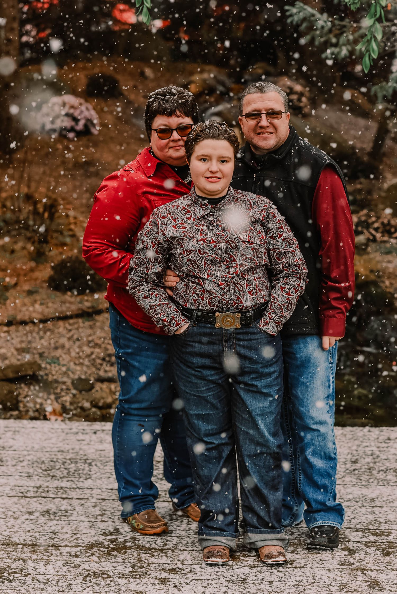 Three people pose together in warm winter clothing while snow falls around them.