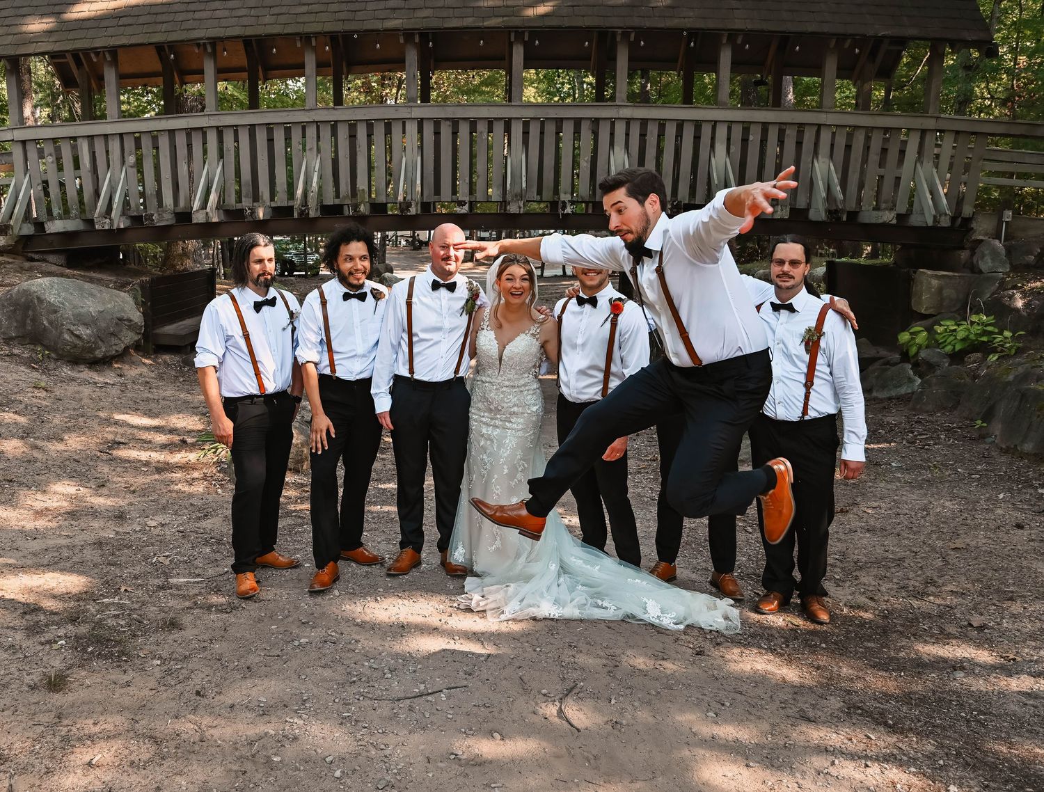 Wedding party poses playfully in front of rustic wooden bridge.