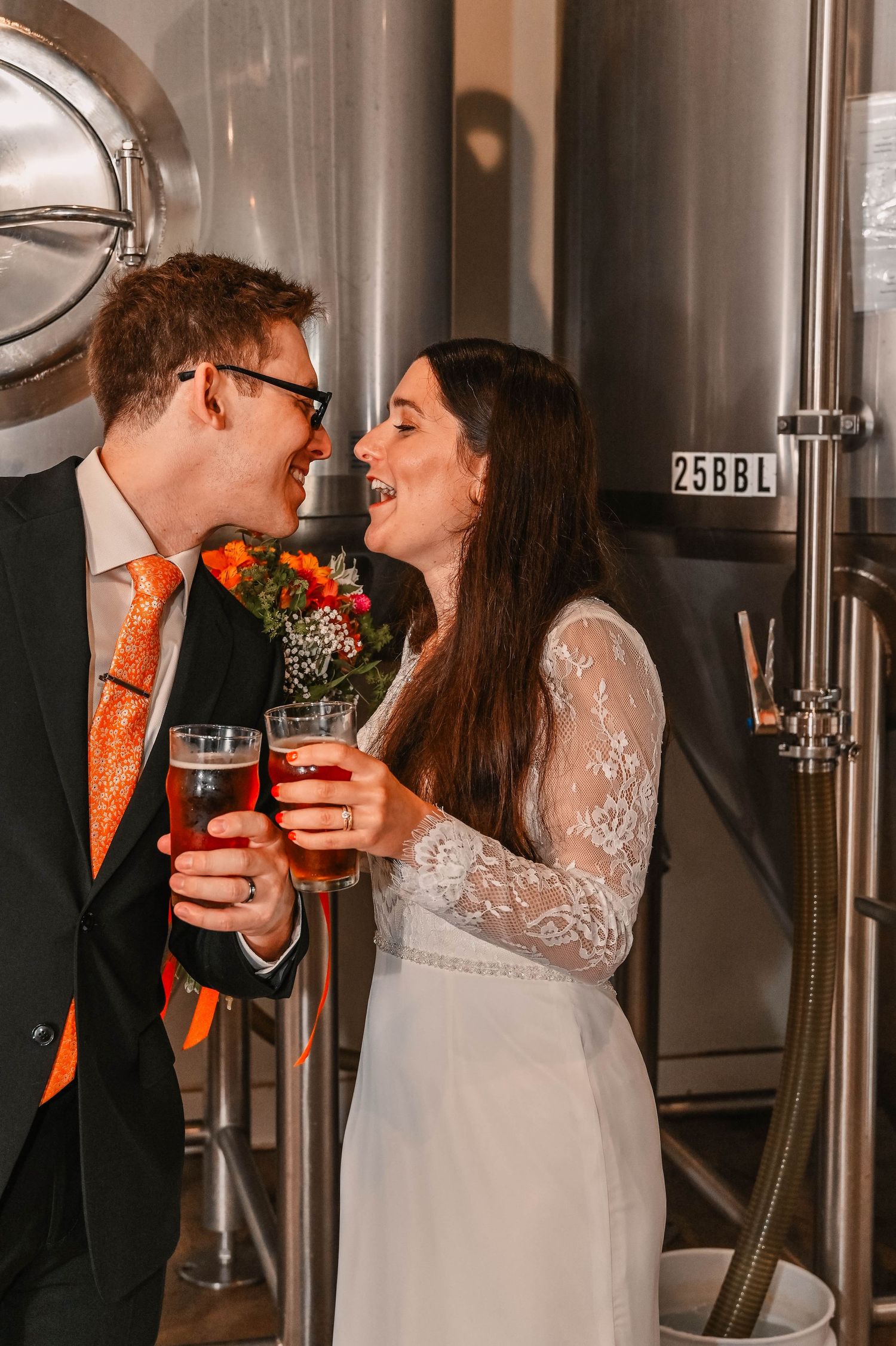 Newlyweds share drinks together in front of large steel brewery tanks.