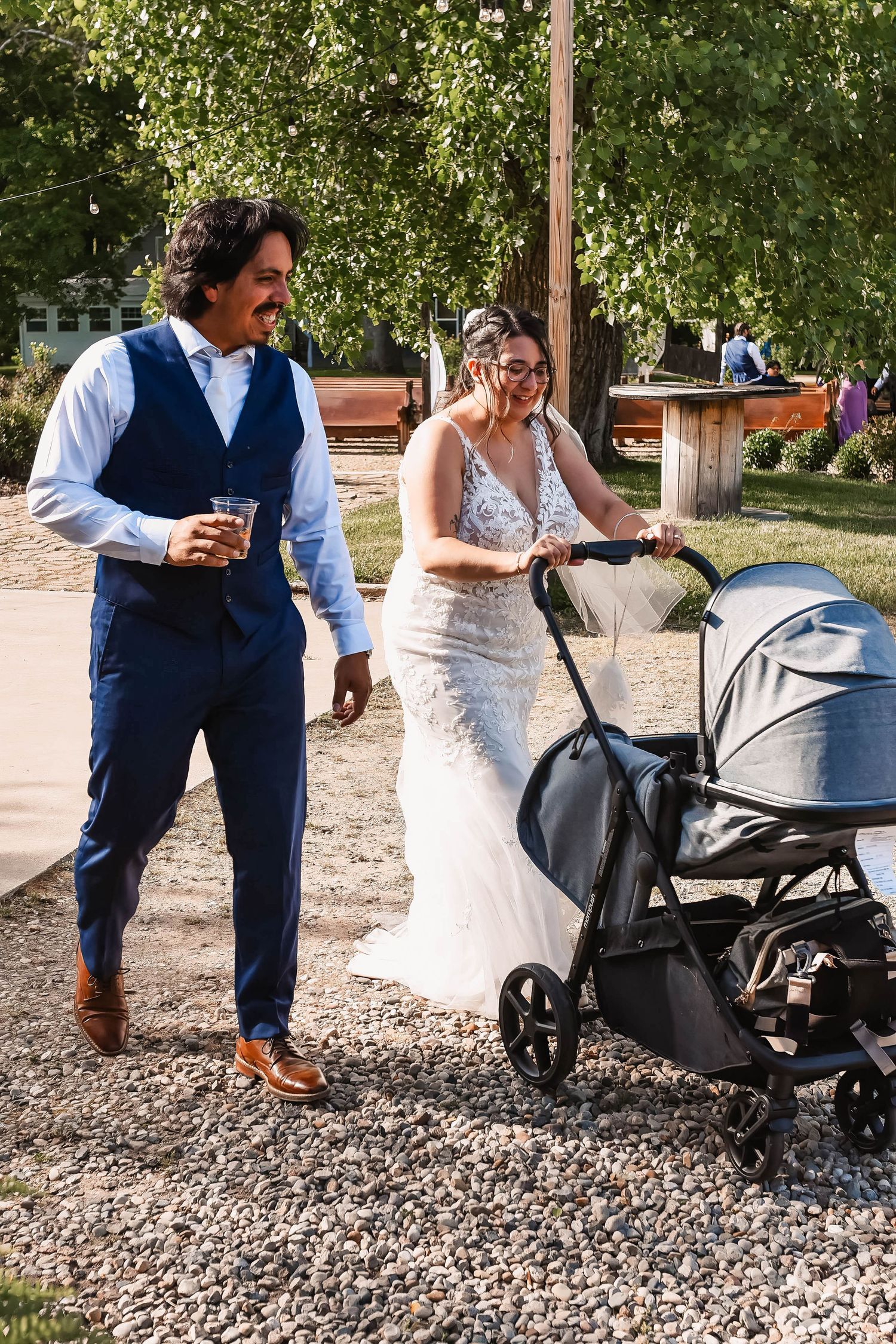 Wedding couple interacts with a baby stroller on a gravel path outdoors.