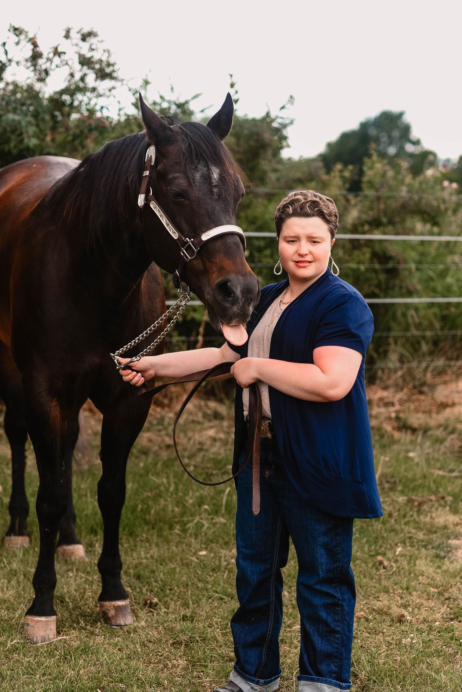 A person in navy blue stands with a dark brown horse in a grassy field.