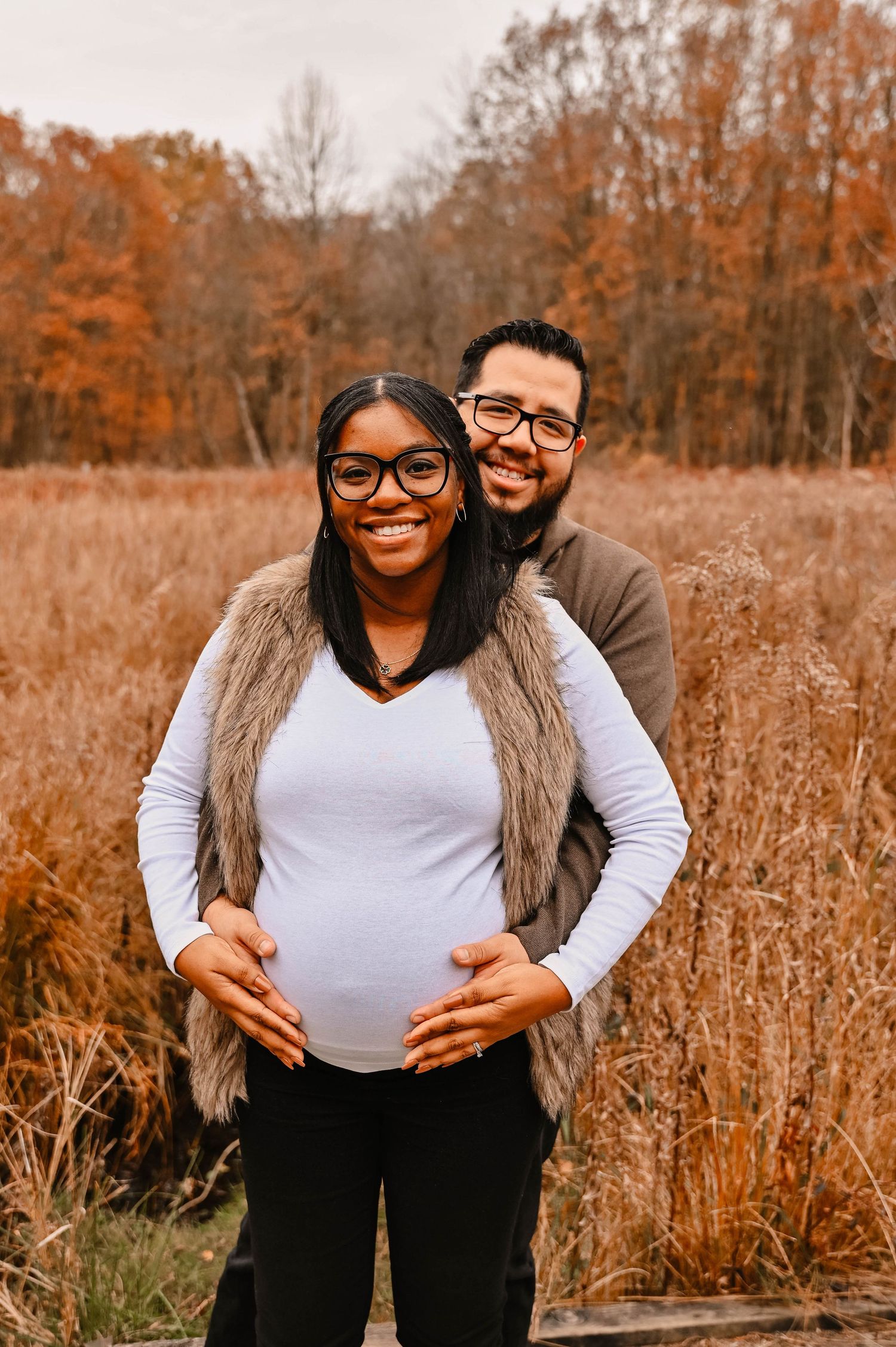 A couple embraces in an autumn field with tall brown grass and trees in the background.