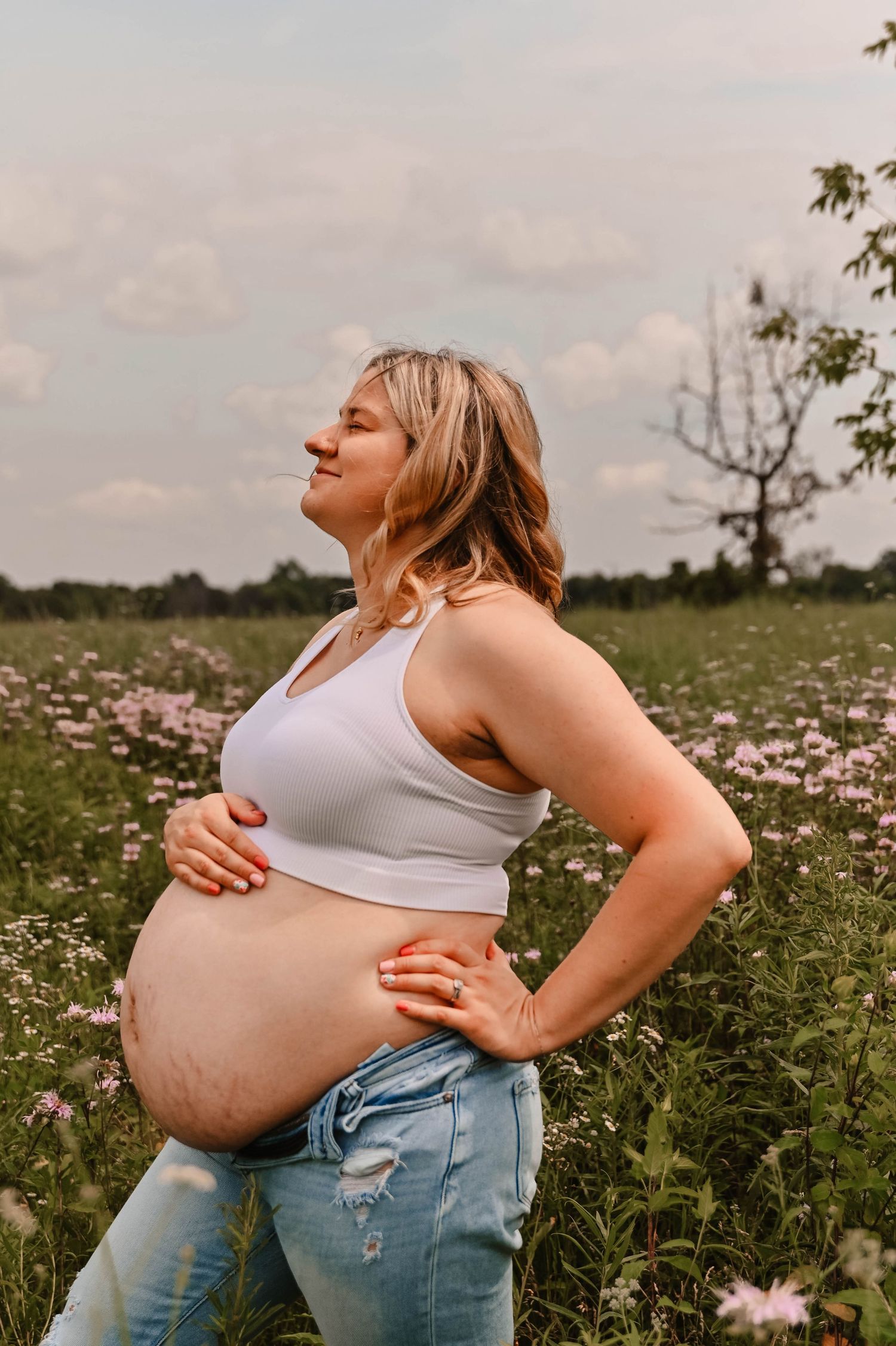 Side profile maternity photos taken in a meadow with pink flowers and cloudy skies.