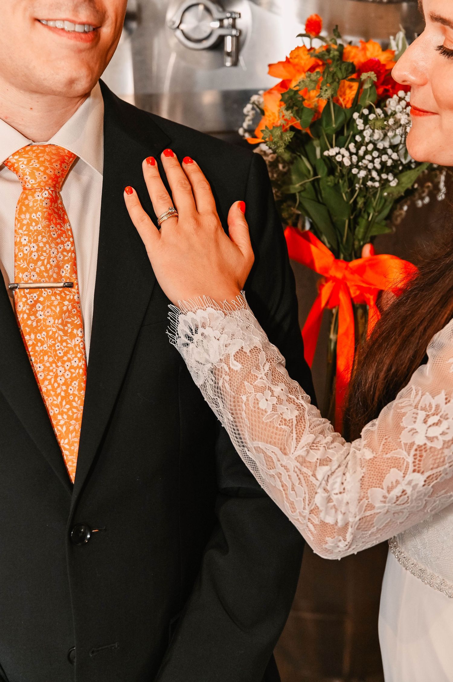 Close-up of orange floral tie and lace sleeve detail with engagement ring and bouquet.