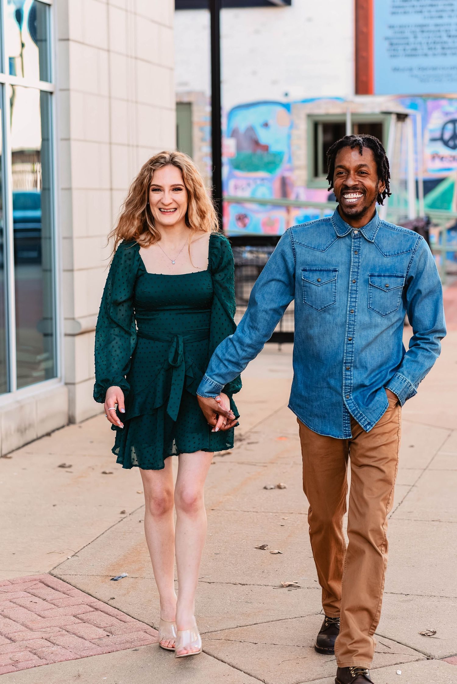 A couple walking hand in hand along a sidewalk wearing denim and dark clothing.