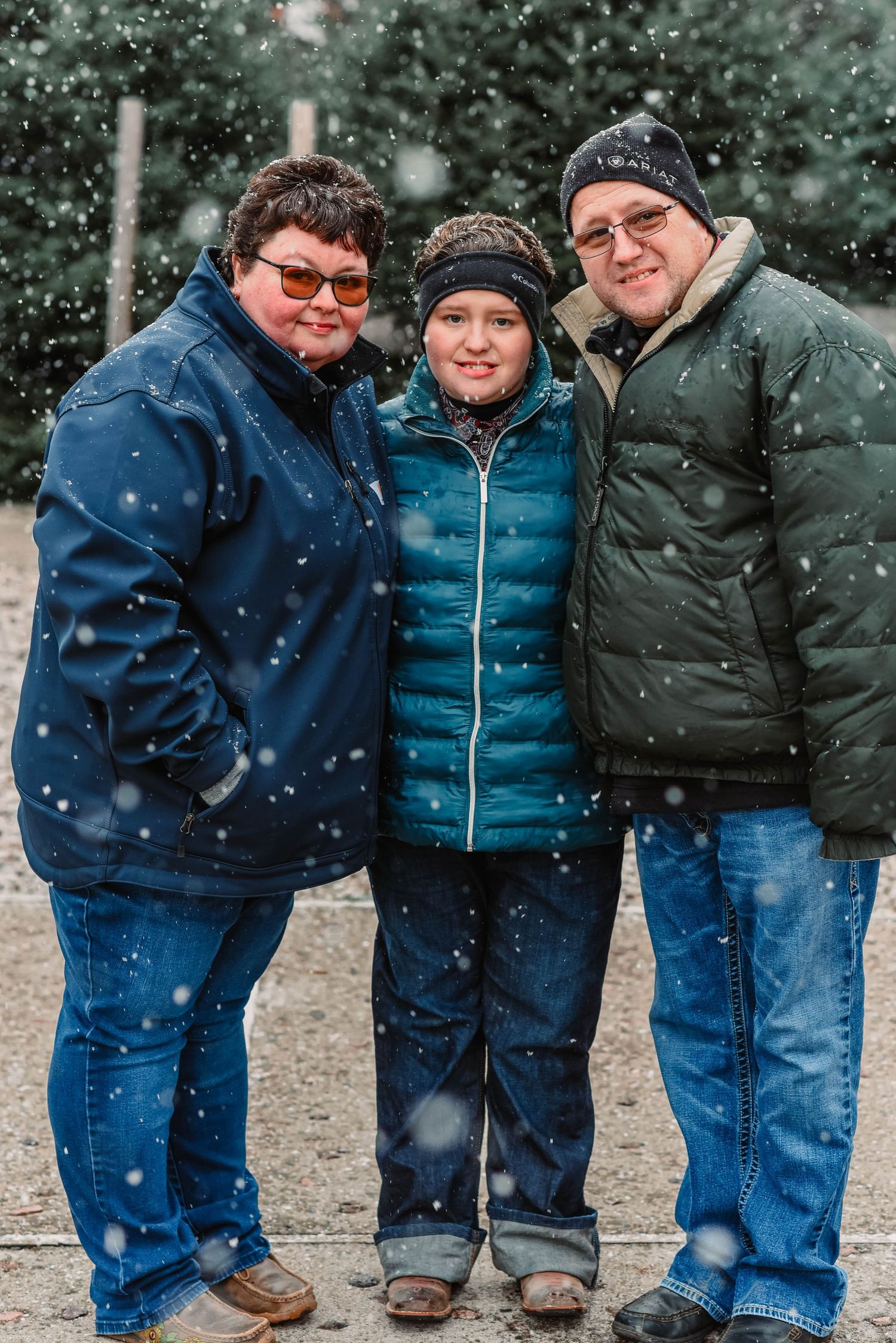 Three people in winter coats smile together as snow falls in the background.