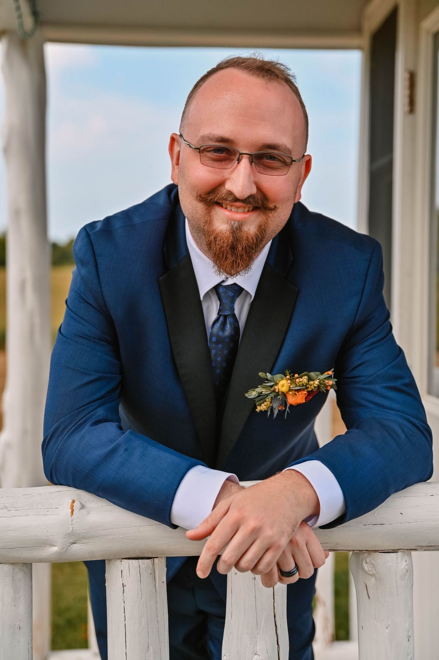 Groomsman in navy suit smiling while leaning on white porch railing.