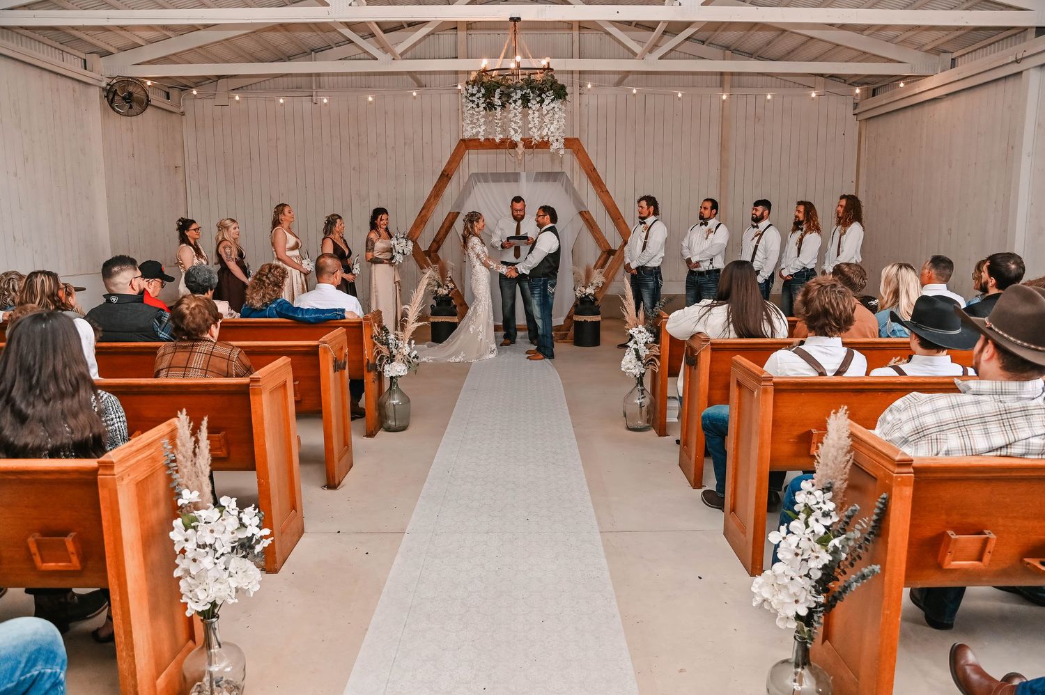 Wedding ceremony in progress in a white chapel with wooden pews decorated with floral arrangements.