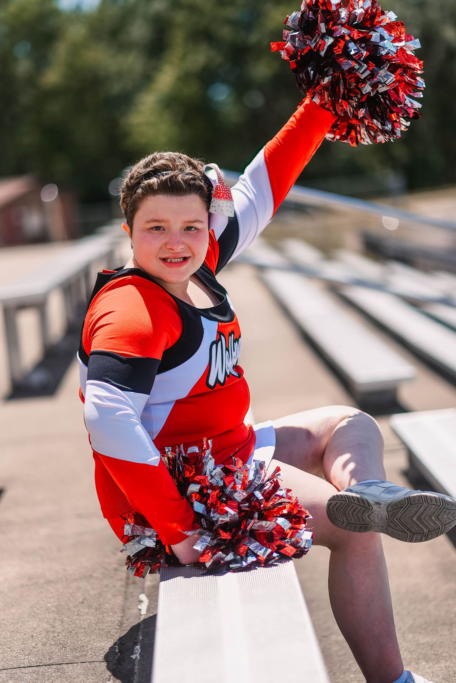 An energetic cheerleader performs with pom poms on stadium bleachers in a red and white uniform.