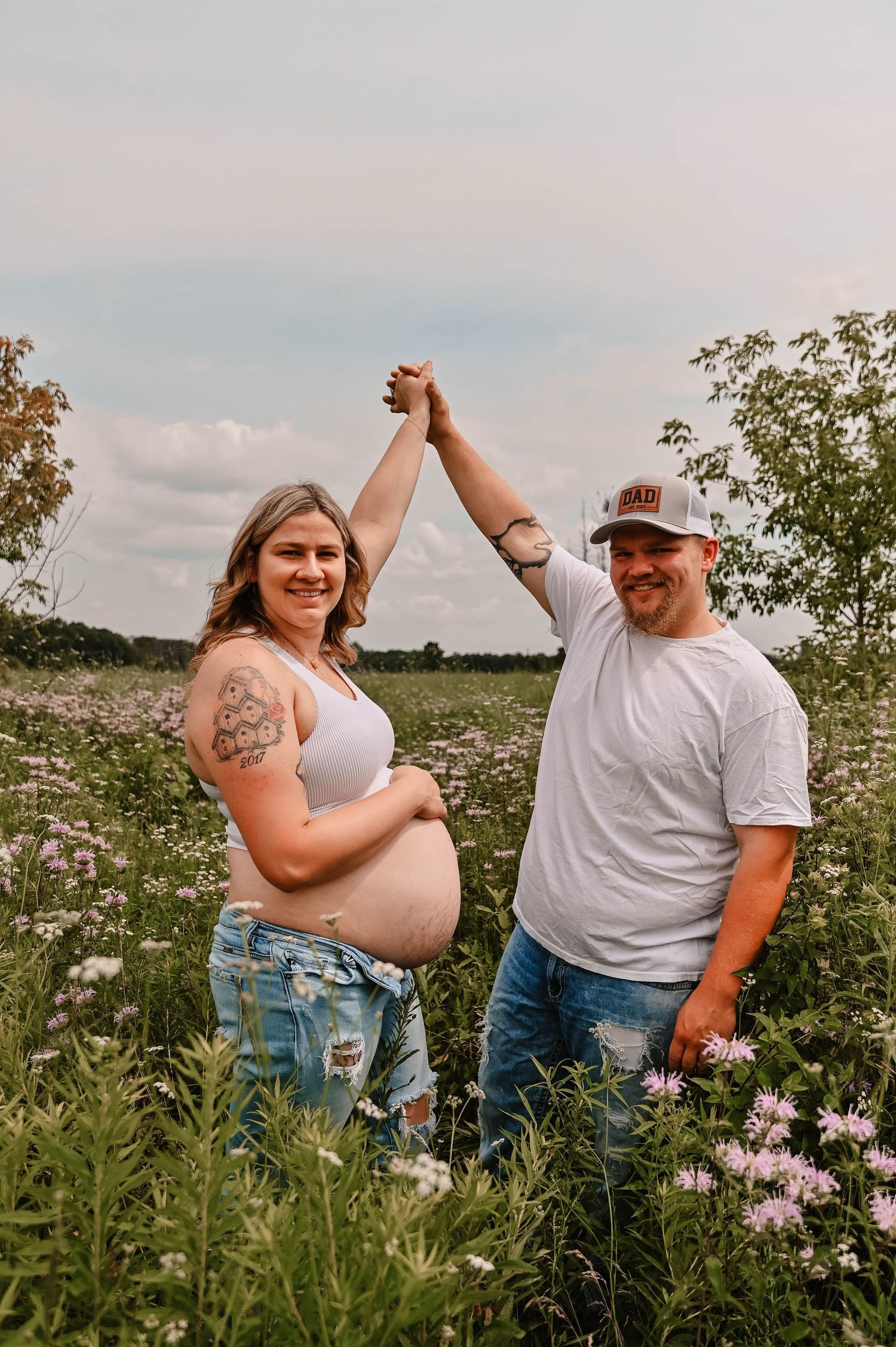 A maternity photoshoot in a wildflower field shows a couple embracing among pink blooms at sunset.