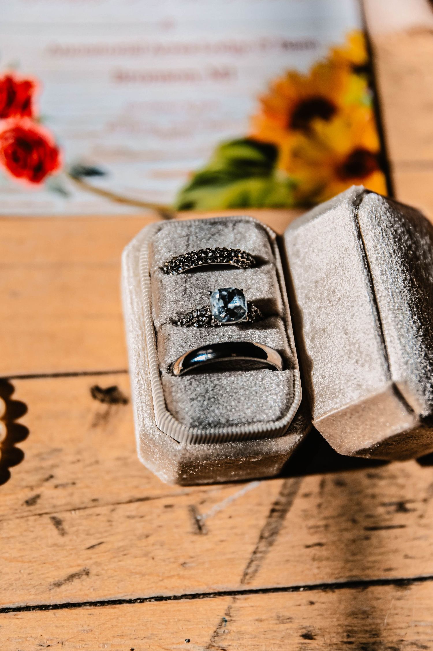 Close-up of engagement ring in grey ring box on wooden surface with sunflowers in background.