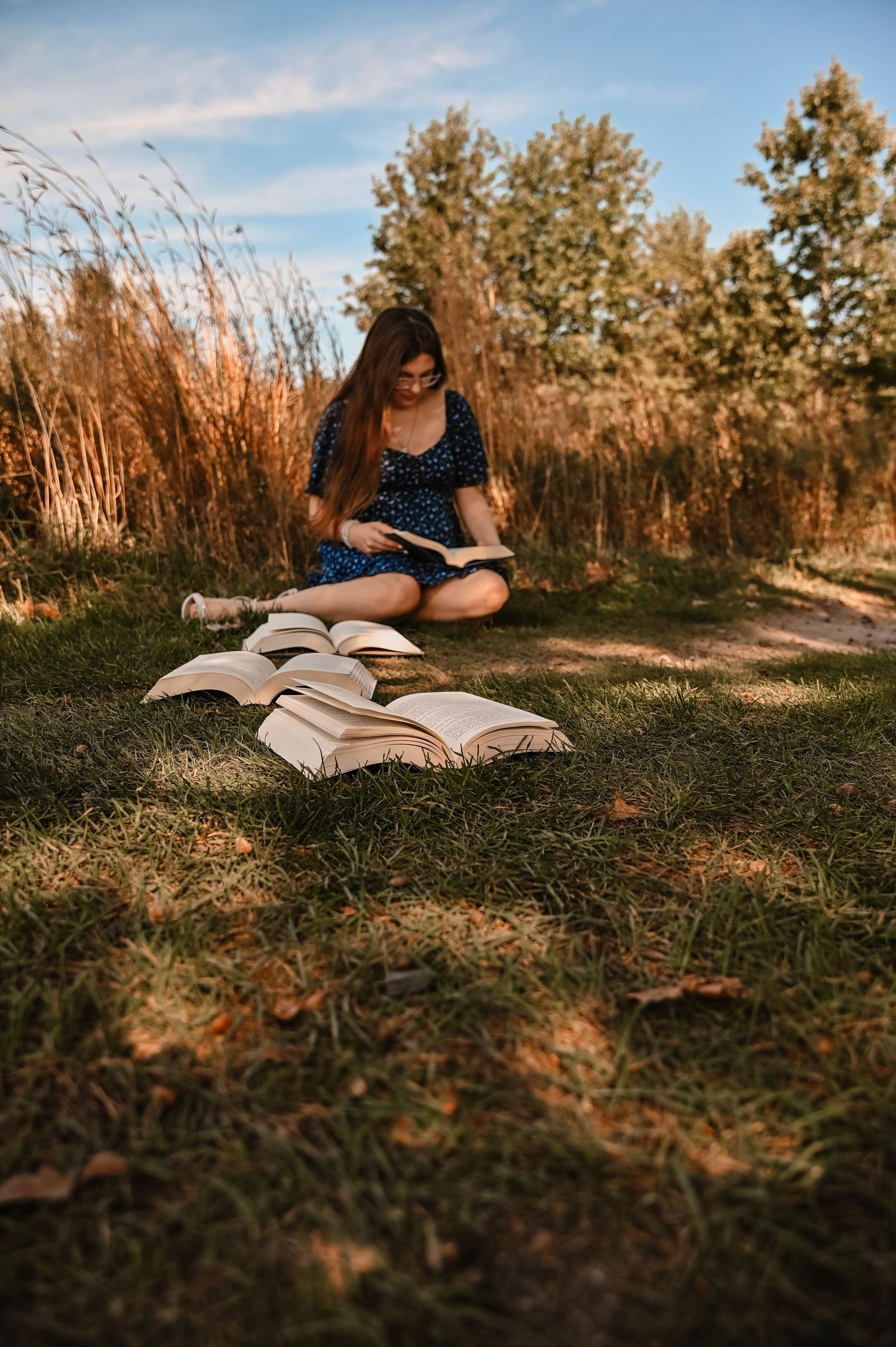 Open books lay on grass while someone in a blue dress reads outdoors on a warm afternoon.