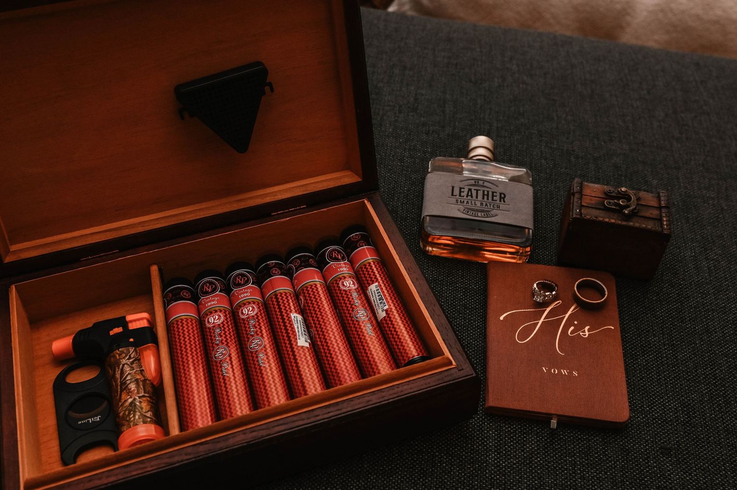 Wedding day accessories including cigars in a wooden box, flask, and rings arranged on dark fabric.