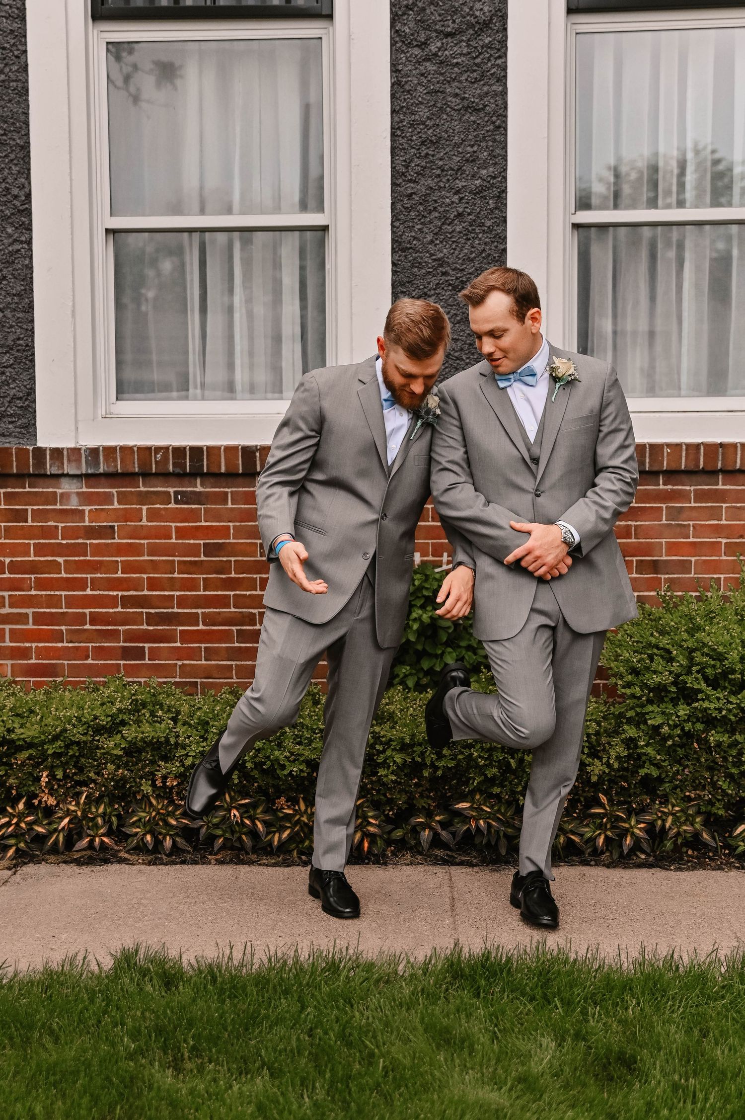 Two groomsmen in matching gray suits perform a playful dance move outside a brick building.