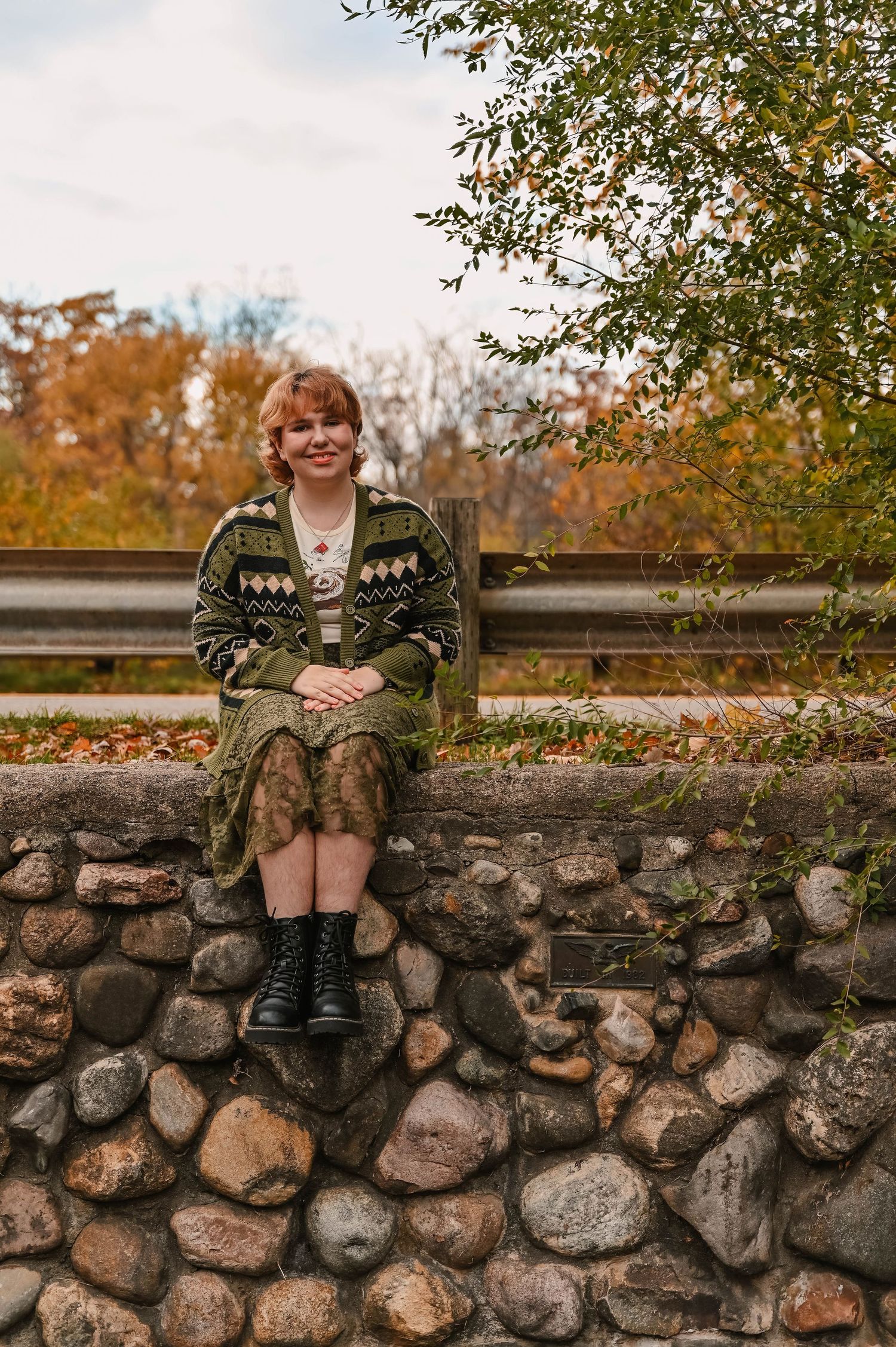 Person in green sweater sits on rustic stone wall during fall season.