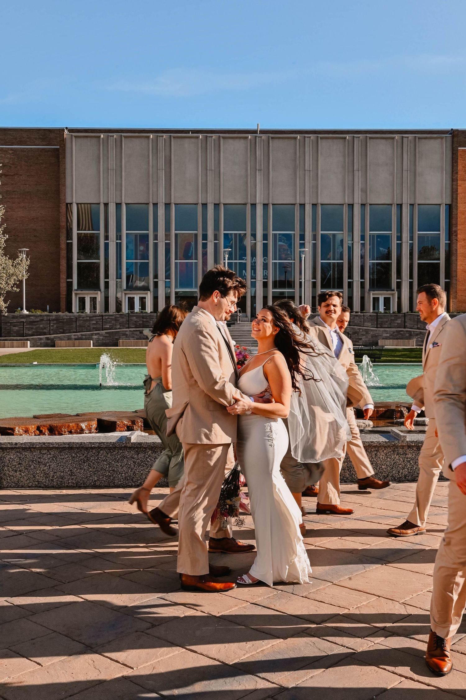 Wedding party in beige and white attire dancing on outdoor patio during daytime celebration.