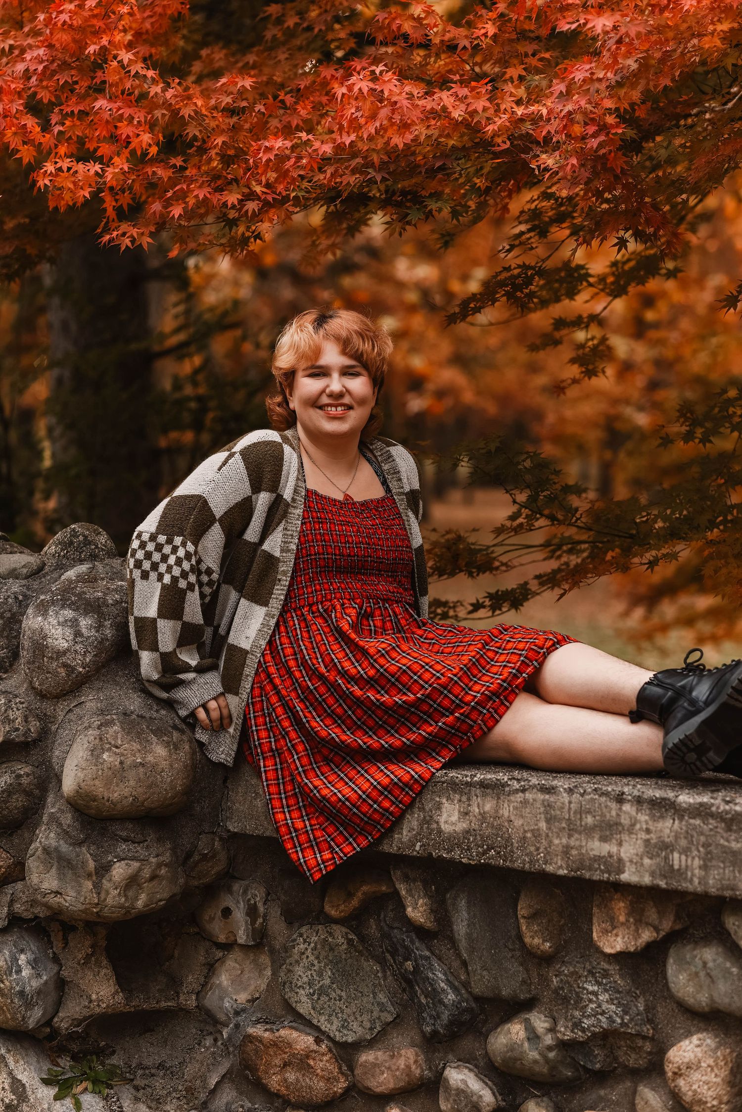 Person in red dress lounges on stone wall surrounded by vibrant autumn foliage.
