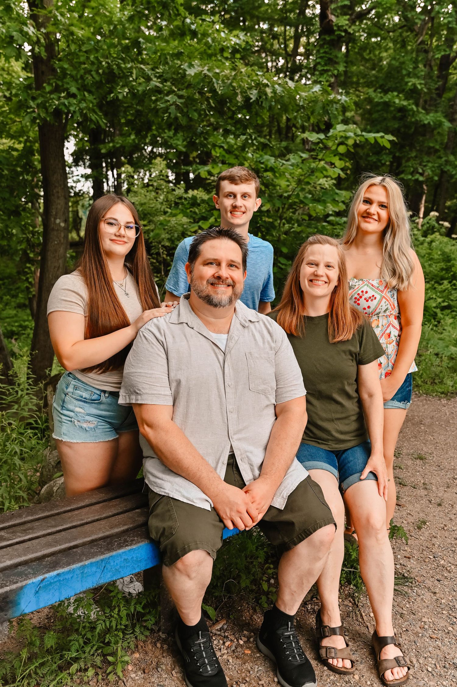 Family members sitting together on a wooden bench in a wooded park setting.