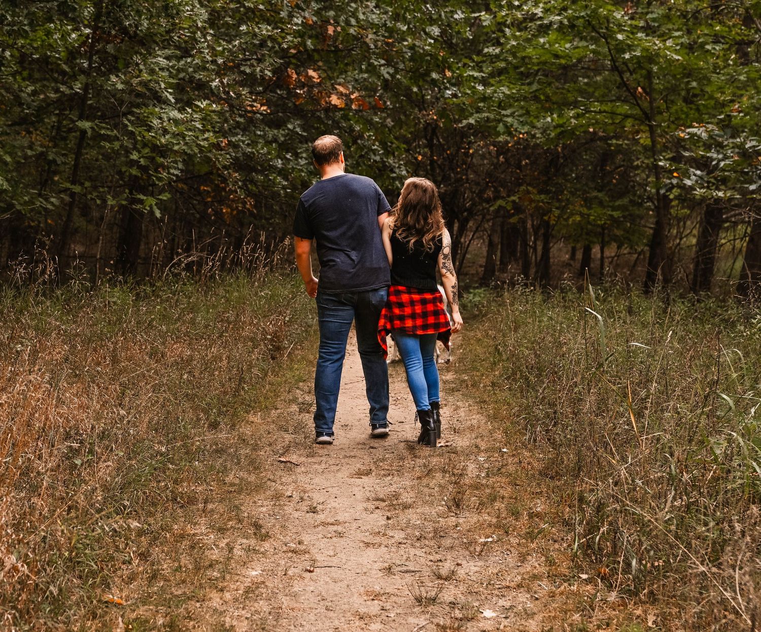 A couple walks hand in hand down a winding dirt path through autumn woods.