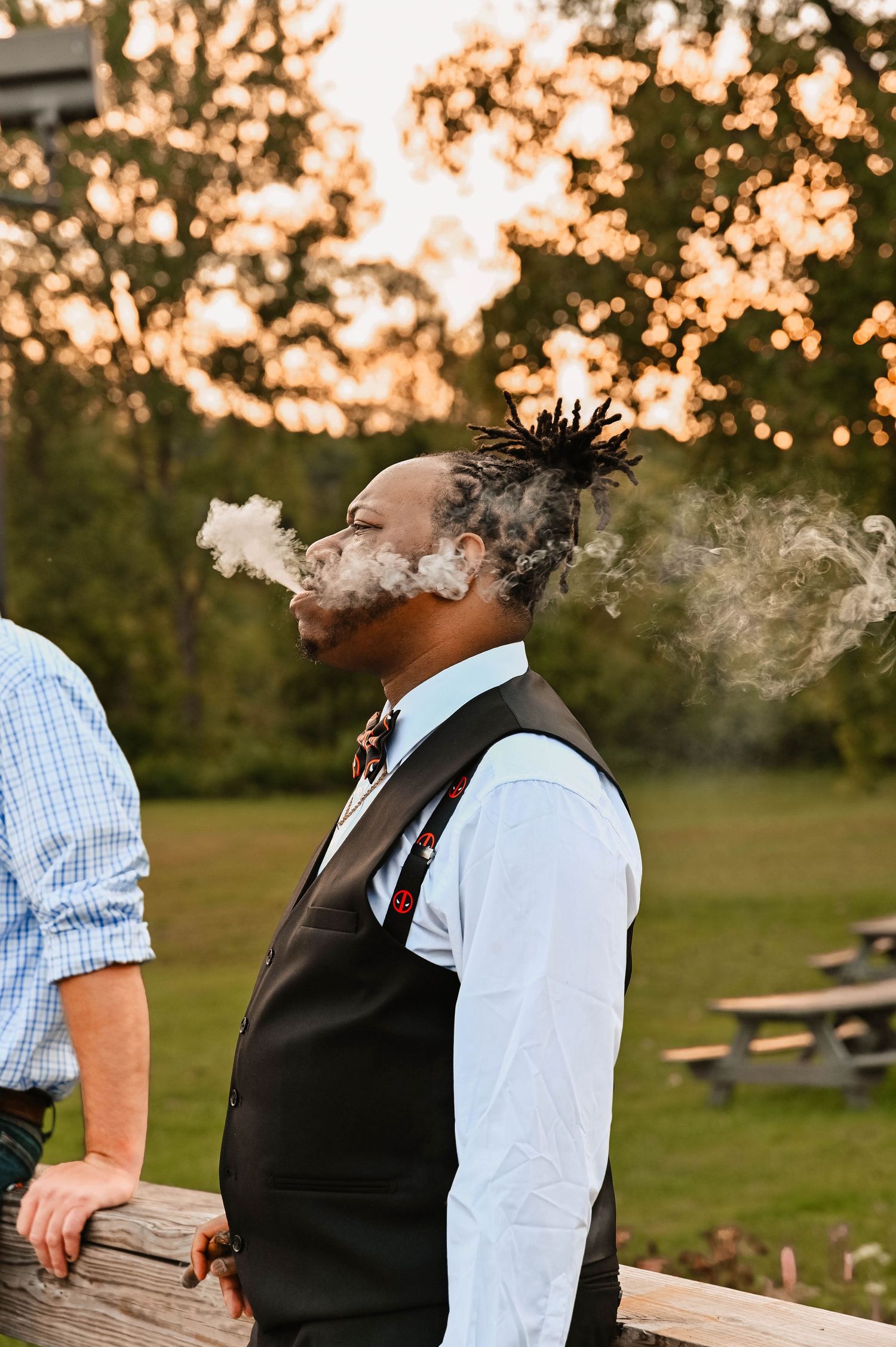 Wedding party member exhales smoke during sunset outdoor celebration.
