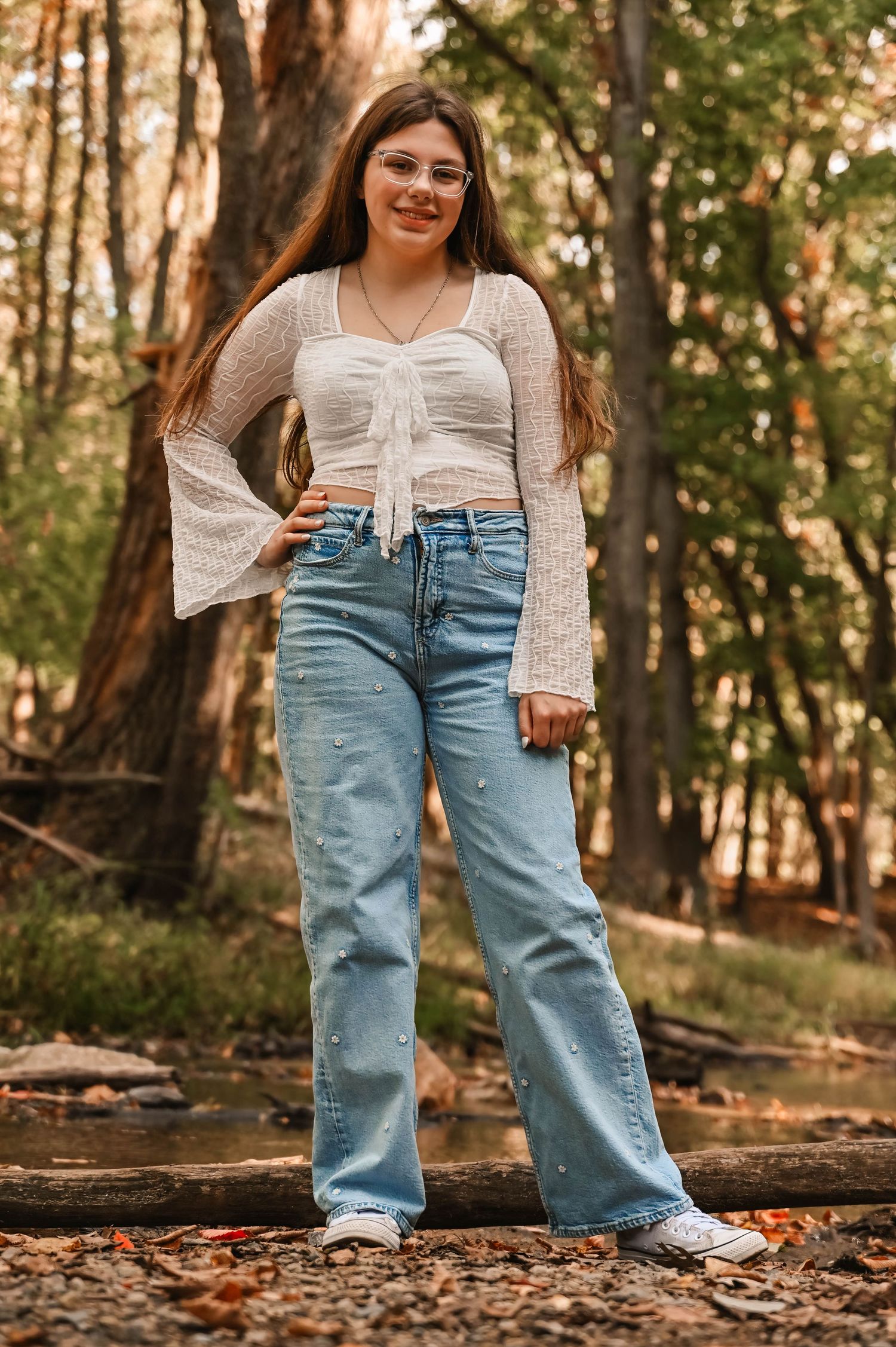 Person in white top and light blue jeans stands among fallen leaves in wooded setting.