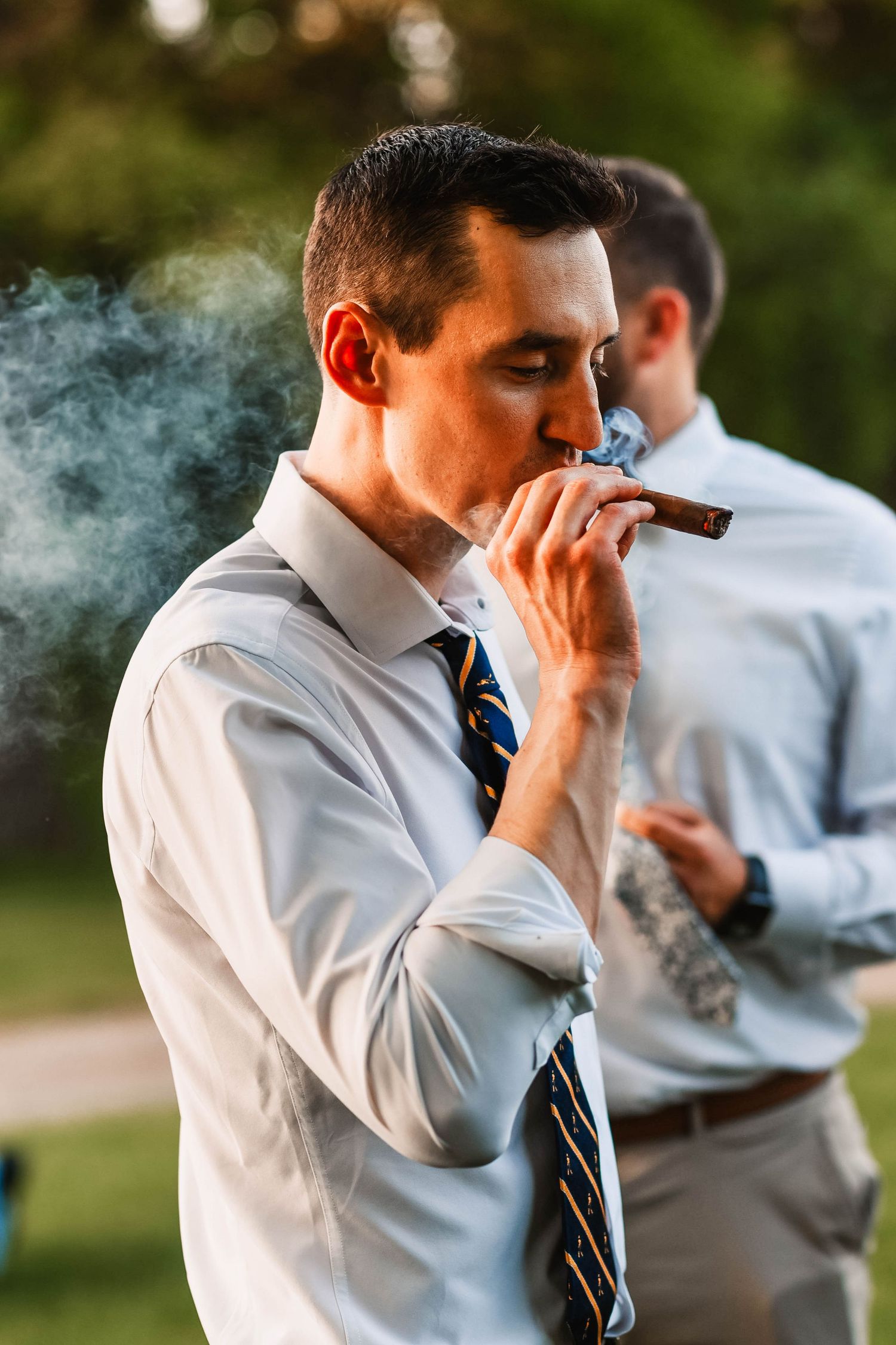 Smoke rises as groomsmen enjoy cigars during outdoor wedding celebration.